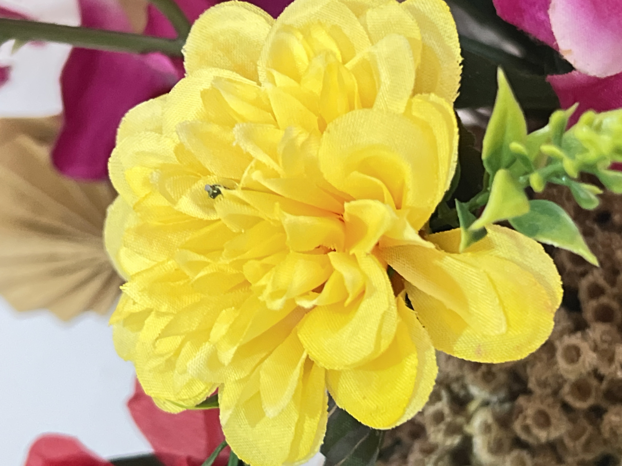 A close-up, eye-level, full shot shows a bright yellow, artificial flower in the foreground. The flower has numerous layered petals with a slightly ruffled texture. A tiny, dark insect, possibly a fly, is perched on one of the central petals. In the background, out-of-focus elements include hints of red and magenta flowers, green foliage, and a tan, fan-shaped object. The lighting is bright, casting soft shadows. The scene does not provide any contextual information that can be used to determine its location within Denpasar, Indonesia.