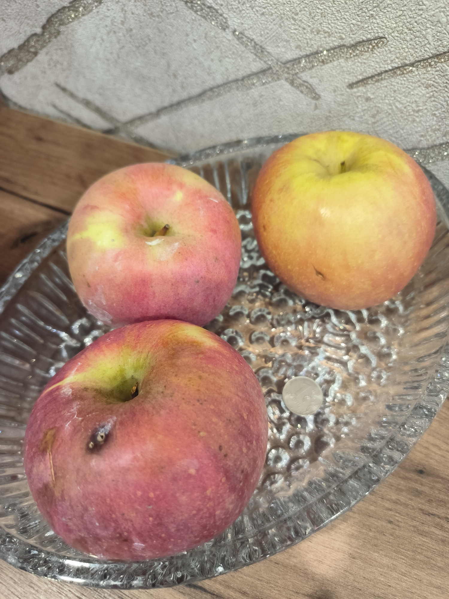Three apples are arranged in a glass dish on a wooden surface. The dish is transparent and has a textured pattern. The apples are a mix of red and yellow. In the dish, there is also a single coin. The background shows a textured wall. This scene is not located in Yuzhno-Sakhalinsk, Russia.