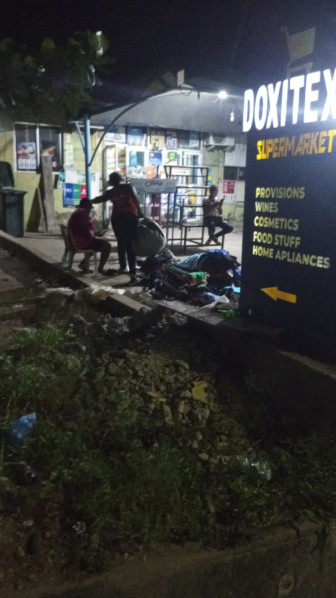 The image captures a nighttime scene outside "DOXITEX SUPERMARKET" in Adankolo, Nigeria, illuminated by the establishment's artificial lights.
Three individuals are present. One person, wearing a red shirt, is seated on a white plastic chair on the pavement on the left side of the frame. Nearby, another person in a darker top stands, leaning over a large pile of items, likely clothes or textiles, spread on the ground near the supermarket sign. A third person is partially visible further back near the supermarket entrance, appearing to be standing or seated by a metal stand.
The prominent feature is the brightly lit sign for "DOXITEX SUPERMARKET," featuring white and yellow lettering. Below the main name, a list of goods is advertised: "PROVISIONS," "WINES," "COSMETICS," "FOOD STUFF," and "HOME APLIANCES" (with "appliances" misspelled). A yellow arrow on the sign points to the left. Through the supermarket's windows, various products on shelves are visible.
The scene suggests an informal gathering or possibly a street vending operation in front of the supermarket. The immediate foreground consists of a grassy, somewhat unkempt patch of ground and a curb. A black trash bin is visible on the far left. The time of day is clearly night, evidenced by the darkness and the reliance on artificial lighting. The overall atmosphere is casual, typical of a busy commercial area after dark.