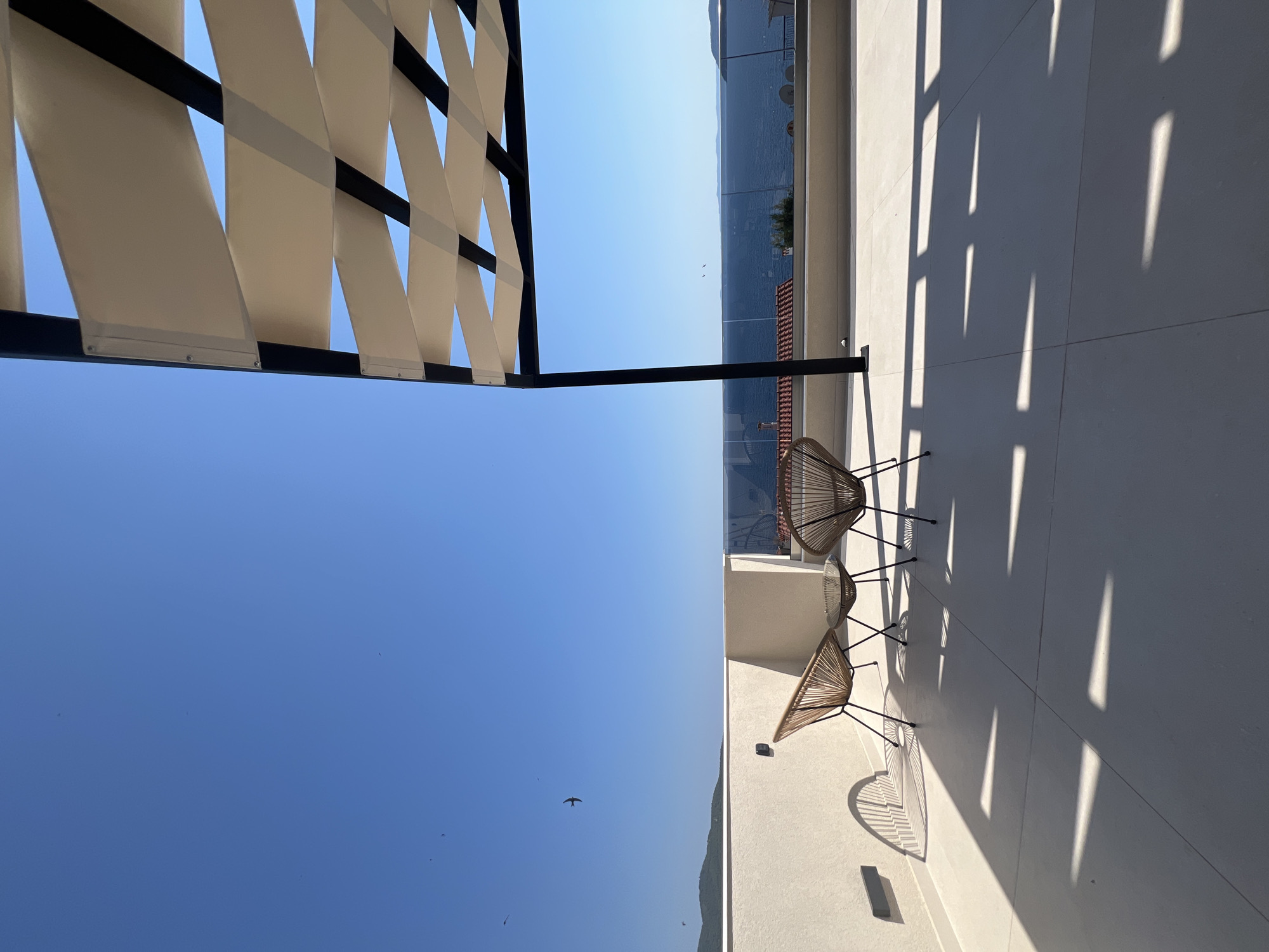 The image depicts a sunny day on a balcony in Sithonia Municipal Unit, Greece. The scene is dominated by a clear blue sky, a glimpse of the sea, and a modern white building. Two wicker chairs with dark metal frames are placed on the balcony, casting distinct shadows on the tiled floor. Above the balcony, a white canopy structure with black metal supports provides shade. The bright sunlight suggests it is daytime, likely late morning or early afternoon, given the length and angle of the shadows. The sea is visible in the background, bordered by landmasses that hint at a coastal or island location. A few birds are seen flying in the distance against the vast blue sky. The overall impression is one of a serene and peaceful outdoor setting, ideal for relaxation. No text is visible in the image.