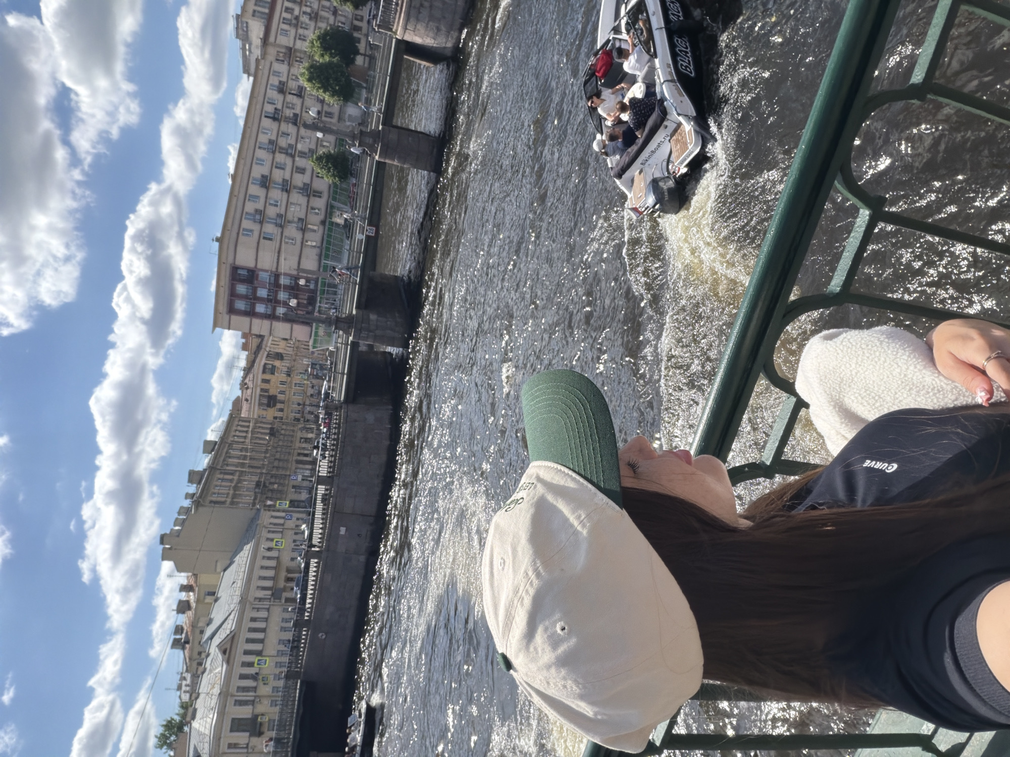 The image captures a sunny day in Saint Petersburg, Russia, with a young woman in the foreground, wearing a light-colored baseball cap with a green brim and a dark t-shirt. She is leaning on a green railing, looking out at a wide canal.
In the canal, several boats are visible. A motorboat, with a white hull and the words "BikiniBoats.ru" and "BLAC ACH" on its side, carries several passengers, all appearing relaxed. Another smaller boat is partially visible further in the distance.
The canal is lined with classic Saint Petersburg architecture. Buildings with light-colored facades, some with ornate details and balconies, stretch along the waterfront. A stone bridge with arched entrances spans the canal in the background, with people visible on it and on the walkways. The sky is a bright blue, dotted with fluffy white clouds, indicating fair weather. The water of the canal is choppy, reflecting the sunlight and creating a shimmering effect. The overall scene suggests a lively and picturesque urban environment.