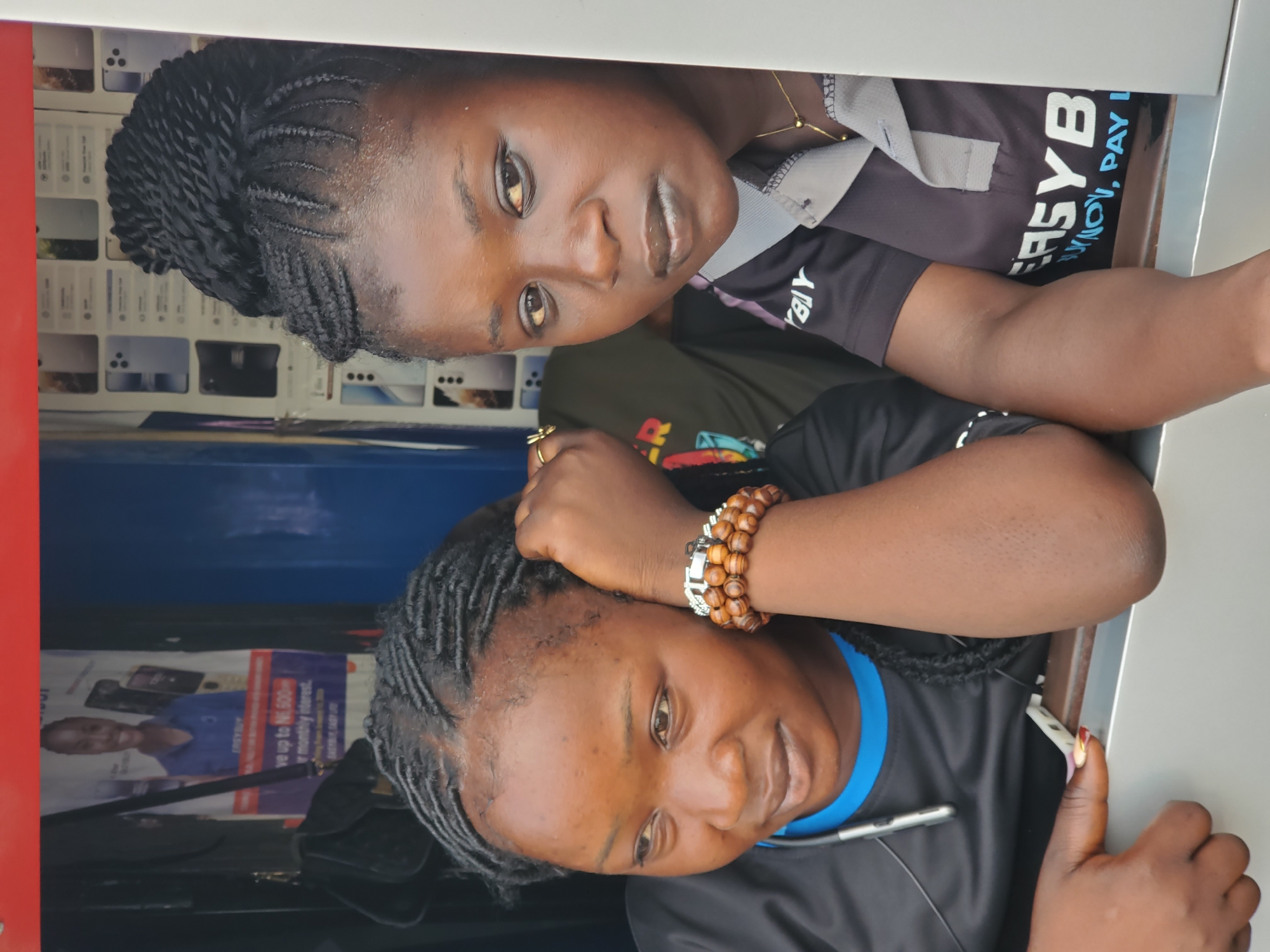 Two young adult women are depicted in an indoor setting, likely a commercial establishment such as an electronics or mobile phone shop, or a service center, in Jos, Nigeria. They are positioned above the viewer, looking down, possibly from behind a counter or partition.
The woman at the top has light brown skin and dark, intricate braided hair. She wears a dark short-sleeved top with a grey collar and white lettering that reads "EASYB" and "S_NOV, PAY I", possibly indicating a service or brand like "EASYBANK" or a payment scheme. She has a thin gold necklace and is looking directly at the camera with a neutral to slightly smiling expression.
The woman positioned below also has light brown skin and dark braided hair. She wears a black top with a blue undershirt visible at the neck. Her left wrist is adorned with a wooden beaded bracelet and a silver watch. Her right hand, featuring colorful fingernails, holds a small white object. She is looking down and to her side with a gentle smile.
The background shows various product displays, possibly mobile phones or their accessories. Behind the bottom woman, a poster advertisement is visible, featuring a smiling woman and text related to a financial offer, including phrases like "up to 16,500-" and "monthly interest". The lighting is bright, suggesting a well-lit interior during the daytime. The overall mood conveyed by their expressions is calm and friendly.