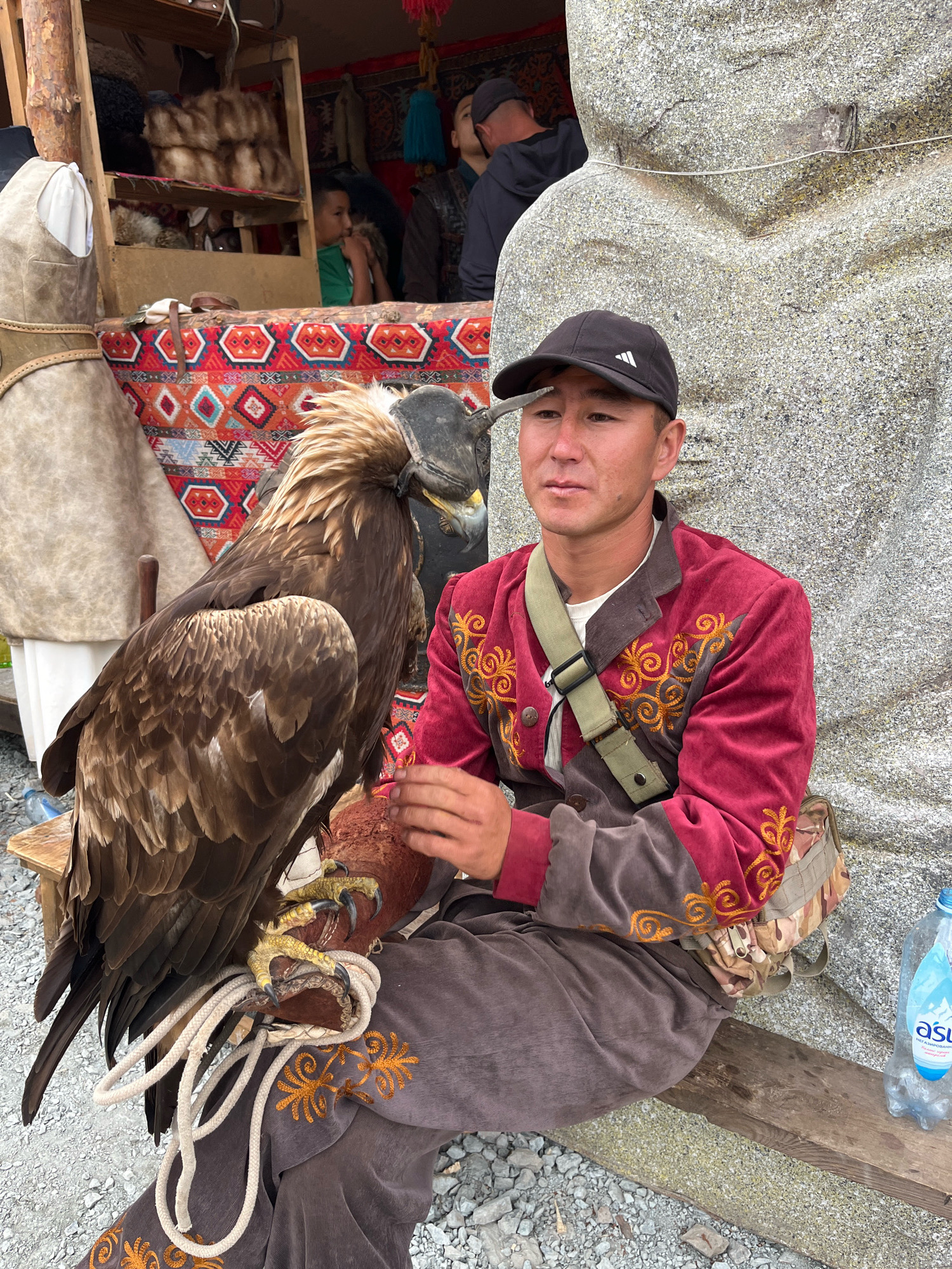 A man in traditional attire sits outdoors with a golden eagle perched on his gloved arm. The man, wearing a dark baseball cap and a red jacket with gold embroidery, looks to his right. The eagle, with its wings spread, has a leather hood over its head. Behind them, a large stone sculpture is visible, along with a colorful textile tapestry and wooden stalls displaying fur items. The ground is covered with gravel. A plastic water bottle is positioned on a wooden plank to the right of the man. The scene appears to be a cultural or tourist attraction in the city of Grigor'yevka, Kyrgyzstan.