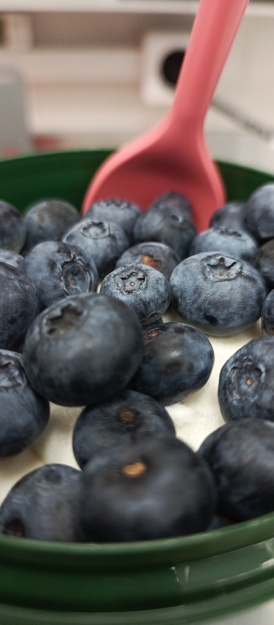 A close-up, high-angle shot captures a pile of fresh blueberries within a green bowl. A pink spoon is partially visible, angled to scoop the berries. The background is blurred, suggesting an indoor setting. The berries appear ripe and are clustered together, with some showing the characteristic dusty bloom. The lighting highlights the texture and deep blue color of the fruit. There is no indication of the location within Bugry, Russia.