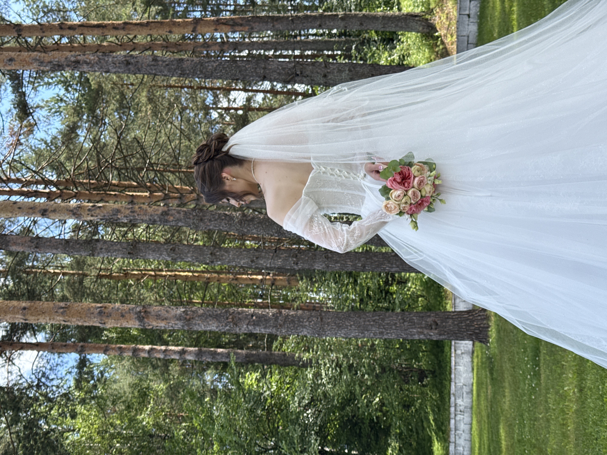 A bride in a white wedding dress with a veil stands facing away from the camera, holding a bouquet of pink and white roses. She is in a forest clearing with tall pine trees and green foliage. The ground is covered in grass with a low stone wall and more greenery in the background. The sky is visible through the trees, indicating an outdoor daytime setting. The scene suggests a wedding ceremony or photoshoot taking place in a natural, wooded environment, likely a park or a secluded area within the city of Votkinsk, Russia.