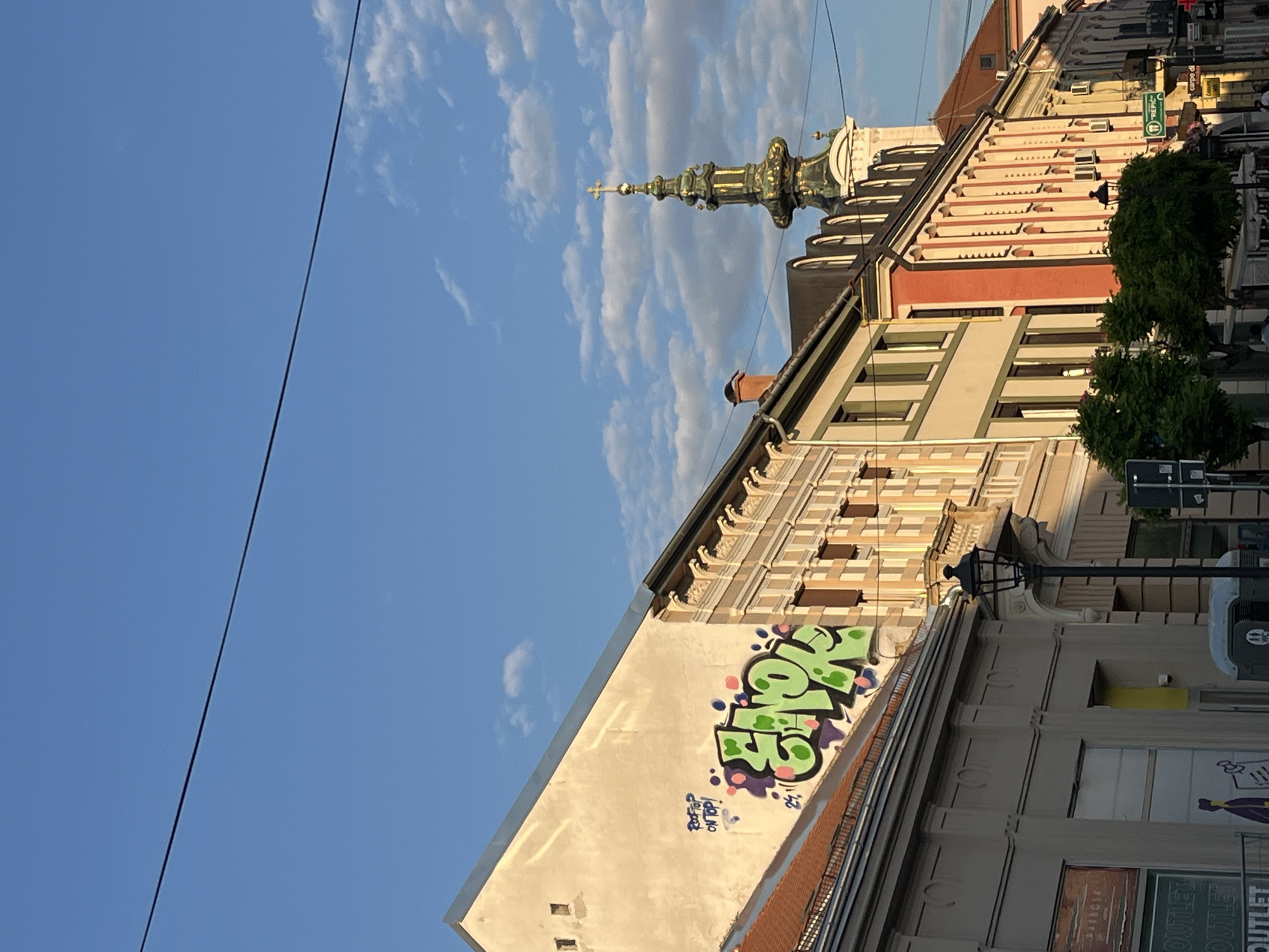 The image shows a street scene in Novi Sad, Serbia. Buildings with varied architecture line the street. The spire of a church is visible. Graffiti adorns one building. The sky is blue with scattered clouds. Trees, utility poles, street signs, and advertisements are present.