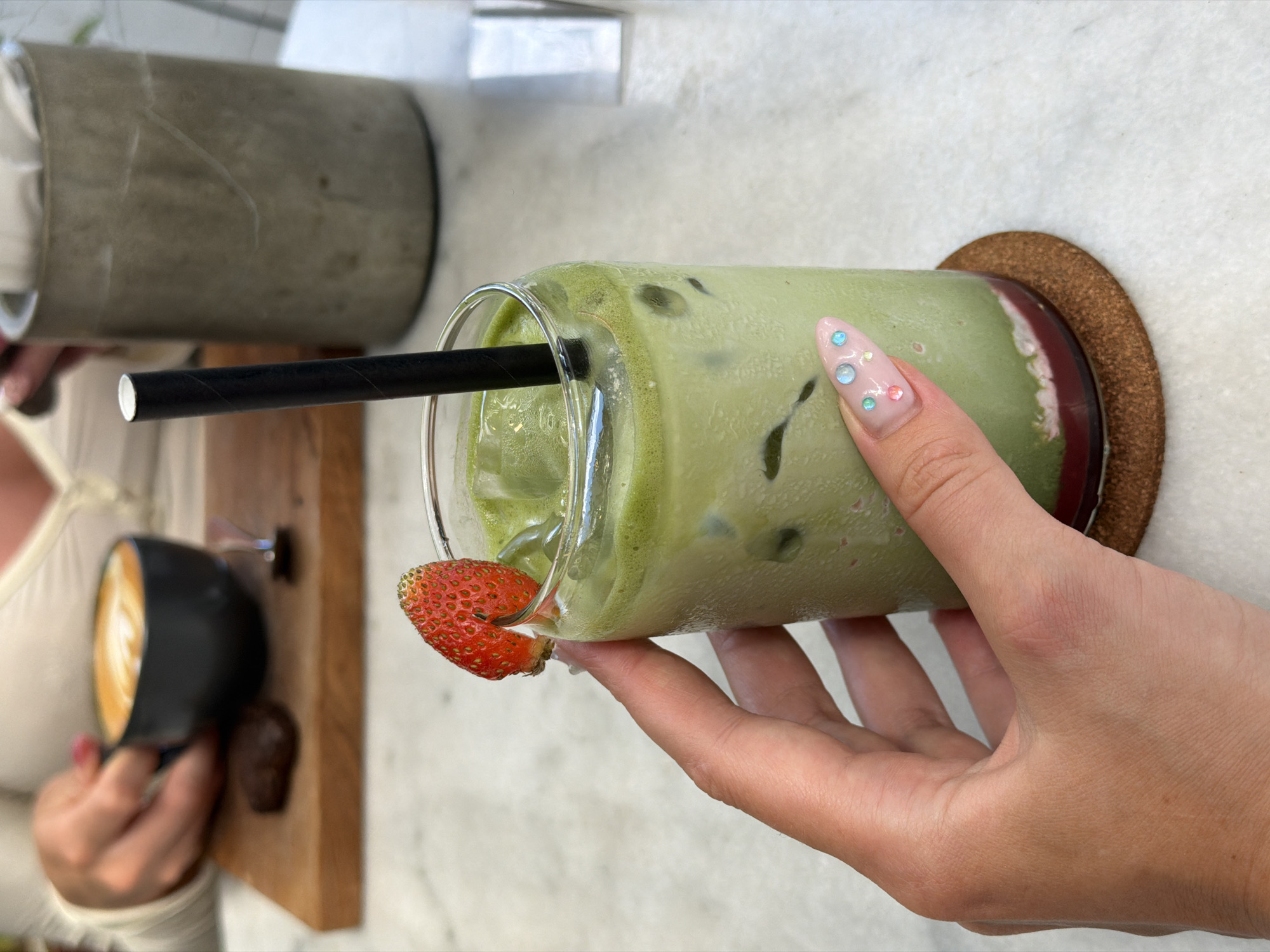 In North Kuta, Indonesia, a hand with light pink nails adorned with small rhinestones holds a clear glass filled with a green iced matcha latte. The drink contains ice cubes and is topped with a slice of strawberry perched on the rim. A black straw extends from the drink. The glass sits on a small, round, brown coaster on a white marble surface. In the background, another person's hands are visible, holding a black cup of coffee with latte art, which rests on a wooden board next to a dark cookie. A concrete cylindrical container with white items inside is also present in the background. The scene appears to be in a cafe or similar establishment.