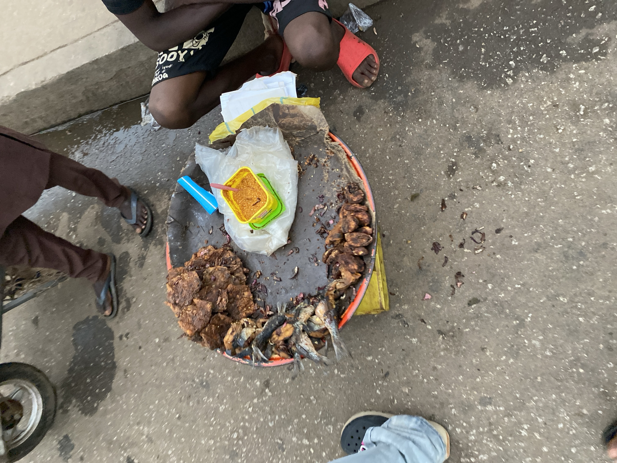 In an outdoor environment, possibly in Bukuru, Nigeria, a vendor is presenting a food tray on the ground. The tray contains assorted foods, including fried meat, fish, and a yellow container of what appears to be spice. People are gathered around the tray; one person wearing brown pants and flip-flops, another person wearing a black t-shirt, shorts, and red sandals, and a third person wearing gray pants and dark shoes. A bicycle wheel is visible on the left. The ground is made of concrete.