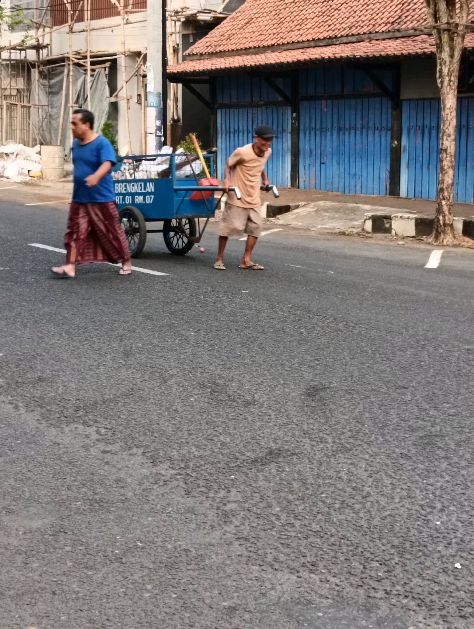 Two men are walking down a street in Purworejo, Indonesia. The man on the left is wearing a blue shirt and a maroon sarong, and is walking next to a blue cart. The man on the right is wearing a tan shirt and tan shorts, and is holding onto the handles of the cart. The cart is filled with items, and has the words "BRENGKELAN RT.01 RW.07" printed on its side. The street is paved and there is a sidewalk on the right side of the road. In the background, there are buildings under construction and a blue building with an orange tiled roof. The time of day appears to be late afternoon or early evening.