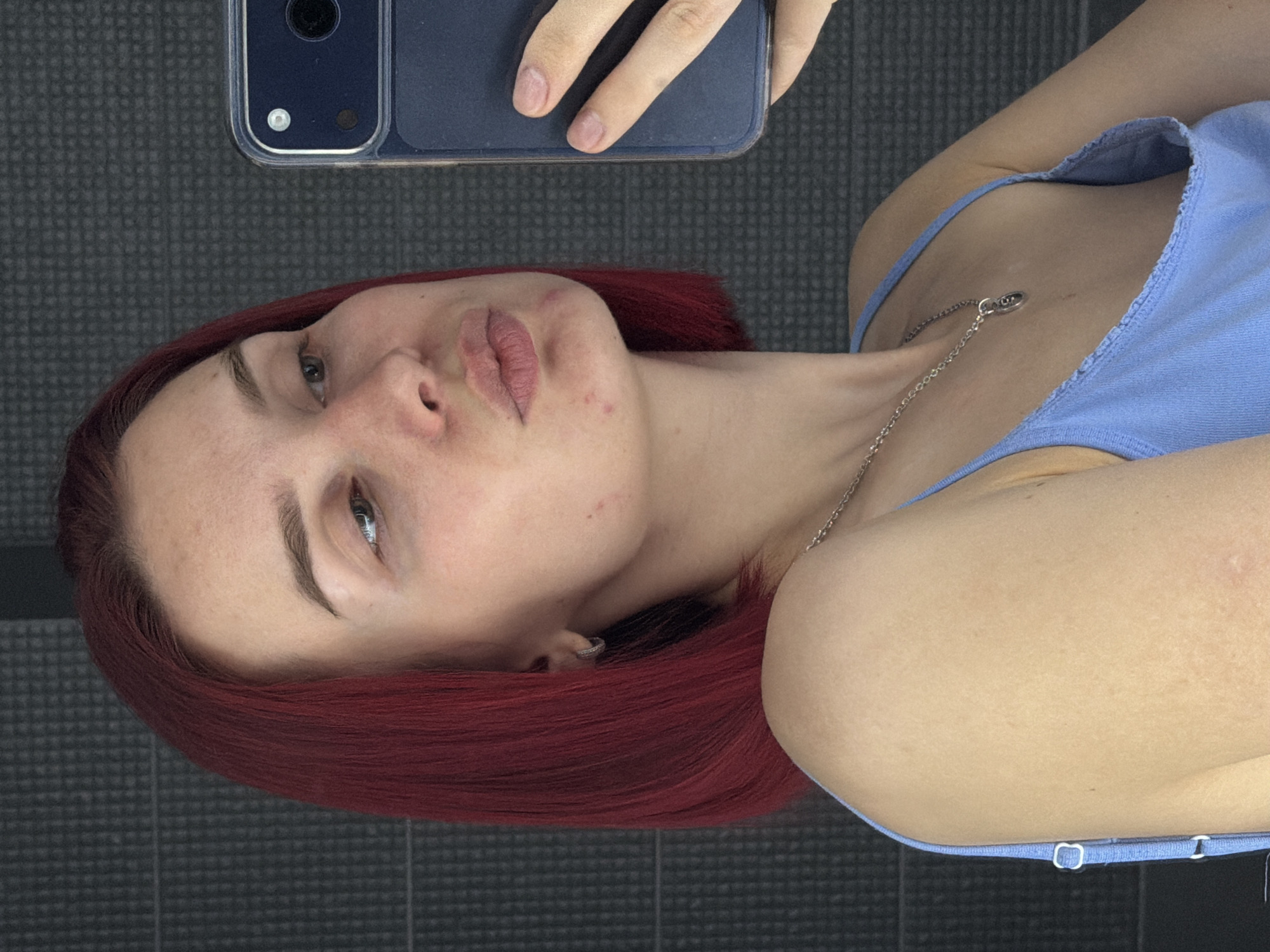 A close-up image, rotated counter-clockwise, depicts a young person with vibrant red/auburn hair taking a selfie while lying down. The subject's head is tilted, and they look towards the camera with a posed expression, including slightly pursed, full lips with a pinkish color and some darker contouring. Their light-colored eyes are open, and dark, well-defined eyebrows frame them. Minor skin blemishes are visible on the cheek and around the mouth. The person wears a light blue camisole or tank top with thin straps, and a silver chain necklace with a small, elongated oval pendant rests on their chest. In the upper portion of the frame, a hand holds a dark-colored smartphone, likely an iPhone given the camera module design, which is partially visible with its dual lenses and flash through a clear case. The background is a dark, textured surface, possibly a floor mat or carpet, featuring a repeating grid pattern. The scene appears to be indoors. No visible text is present in the image.
