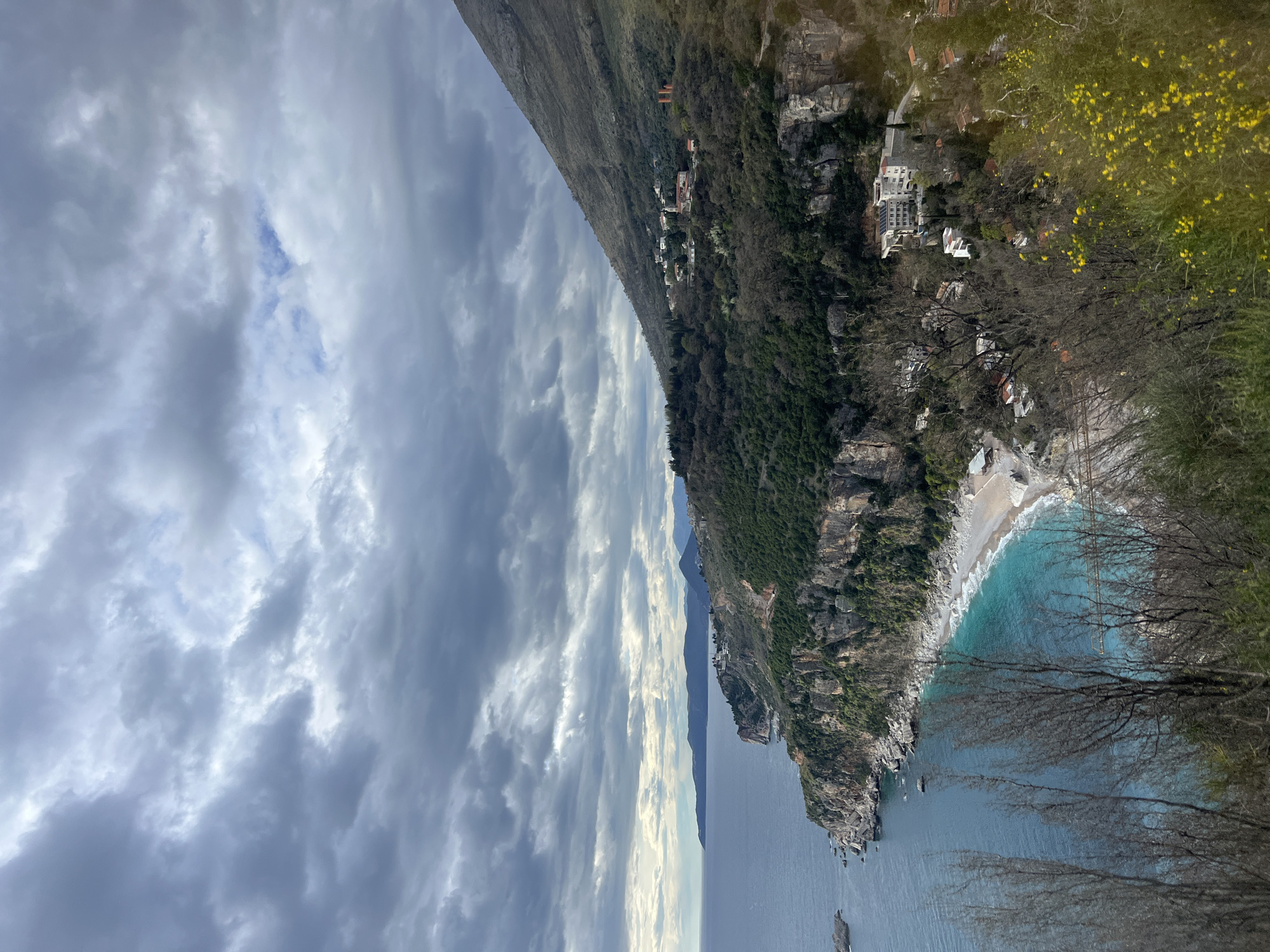 The image is a scenic, slightly rotated, eye-level shot of Perazića Do, Montenegro. The sky is overcast with layered, gray clouds. A mountainous ridge with sparse vegetation and scattered dwellings runs from the upper-left to the mid-right of the frame. A dense, forested cliffside leads down to a narrow beach with turquoise water. Bare tree branches frame the bottom-right corner of the image. The sea stretches from the bottom to the mid-center of the frame, and a few rocky outcrops are visible in the water near the cliff.