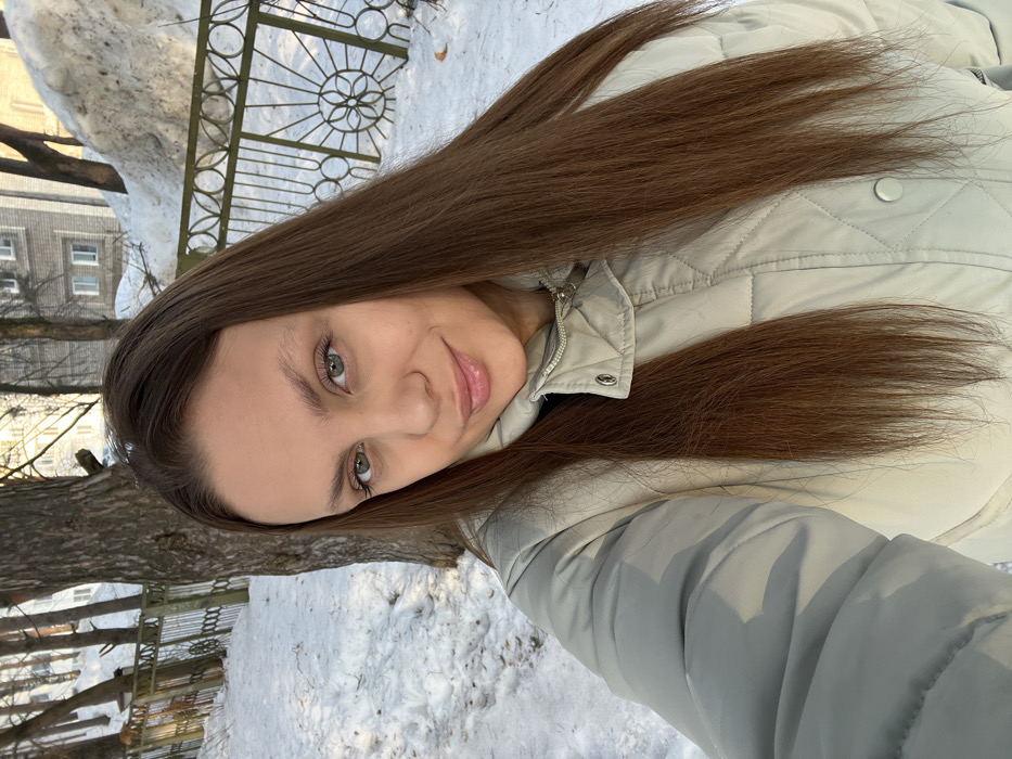 A young adult female, likely in her 20s or early 30s, is depicted in a close-up, selfie-style portrait taken outdoors in Chaikovsky, Russia, during winter. She has long, straight, brown hair and light-colored eyes, exhibiting a subtle closed-lip smile while looking directly at the camera. She wears a light-beige or light-grey quilted puffer jacket, zipped up to her neck. The background features snow-covered ground and several bare-branched trees, with a prominent tree trunk situated directly behind her. A dark-colored ornamental metal fence with circular motifs spans the midground. In the distance, multi-story residential buildings are visible, along with a large mound of snow piled on the right side of the frame. The scene is brightly illuminated by direct sunlight, indicating a clear day.