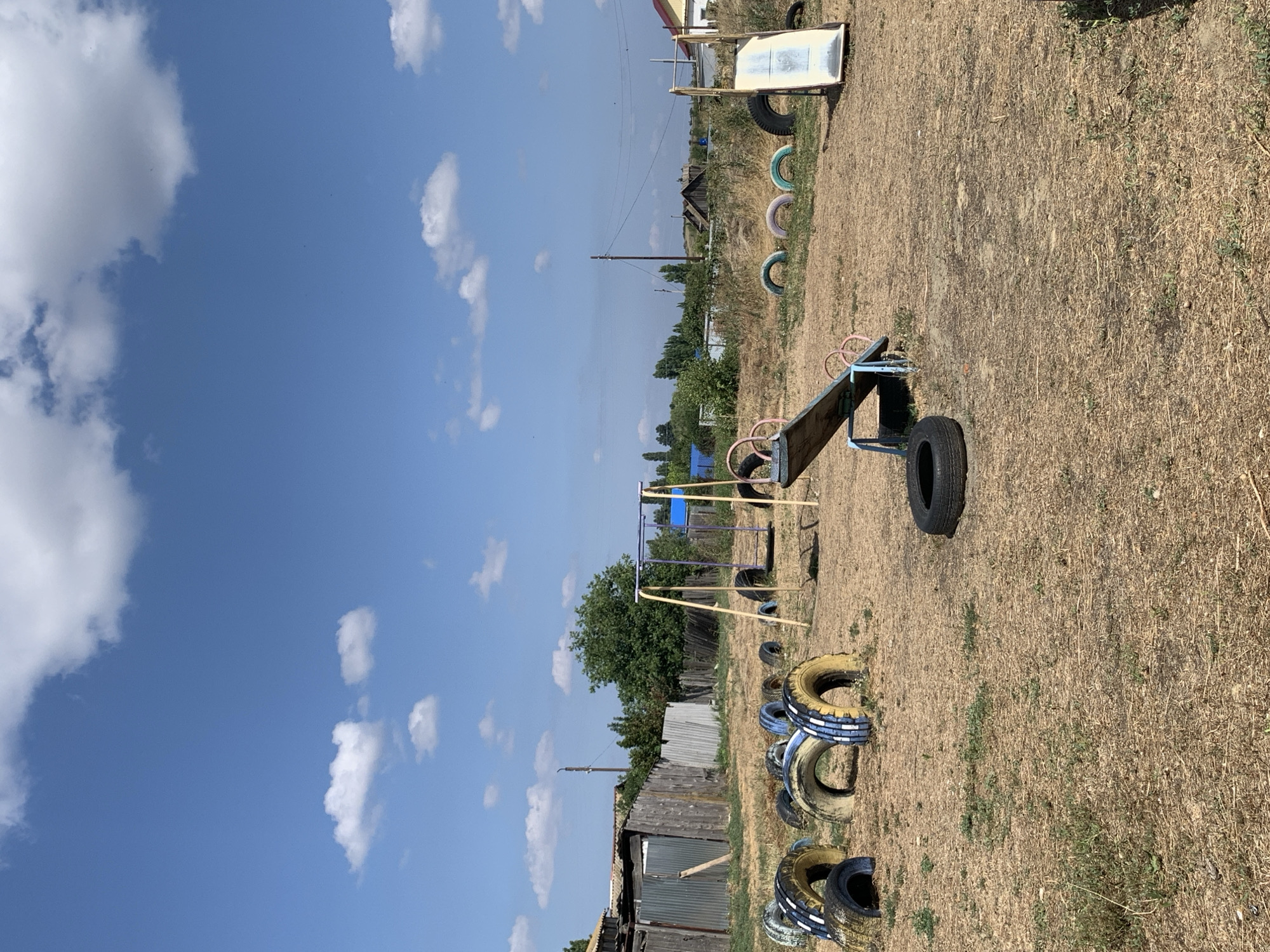 A sunlit, outdoor playground constructed with repurposed tires is depicted under a bright blue sky with scattered white clouds. The playground features a swing set with a blue seat and purple frame, a seesaw with a wooden plank and blue metal supports, and a small slide. Numerous tires, some painted with yellow and blue stripes, are arranged around the area, serving as decorative elements, barriers, and seating. The ground is covered in dry, straw-like grass. In the background, a weathered wooden fence, a metal fence, and various buildings with pitched roofs are visible, suggesting a rural or semi-rural setting. A utility pole with wires stands in the middle ground. The scene appears to be located in Dubovka, Russia, judging by the architectural styles and the dry, continental climate suggested by the vegetation. There are no people or animals present in the image.