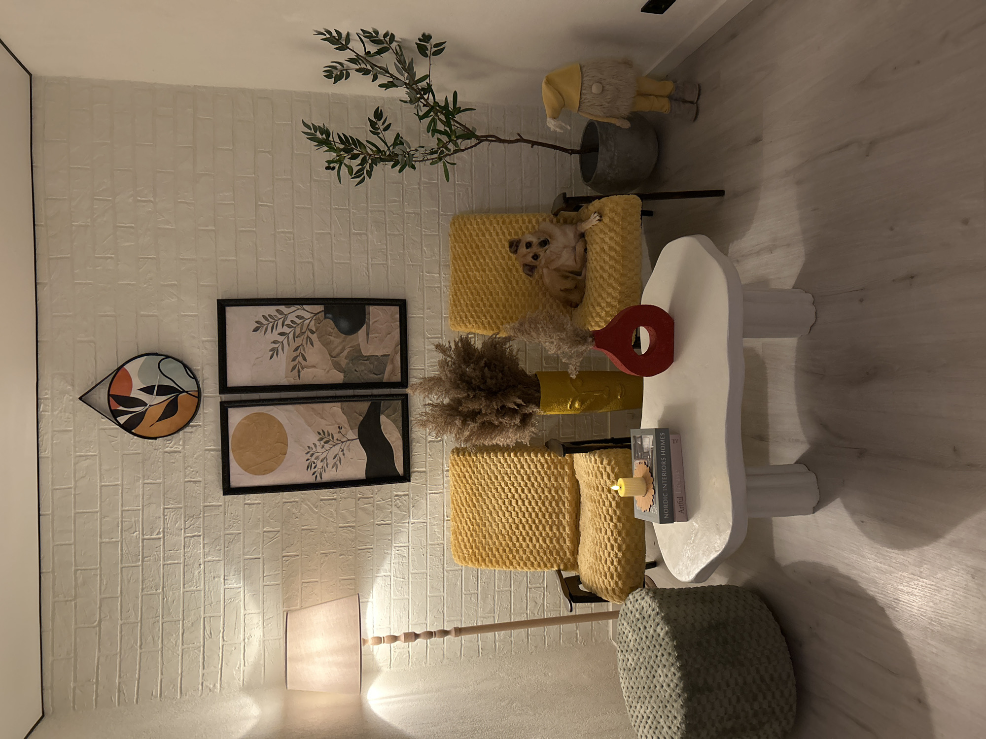 This is a medium shot, eye-level, interior photograph of a living room corner. The walls are white brick, and the floor is light wood laminate.
In the center of the image, two yellow chairs are positioned. The chair on the left is empty, while the chair on the right has a small, light brown dog resting on it, facing the camera. Between the chairs, a white, amorphous-shaped coffee table sits. On the coffee table are two books, a small candle in a brass holder, and a red, ring-shaped vase. Behind the coffee table and between the chairs, a yellow vase filled with dried reeds stands.
To the left of the chairs, a tall floor lamp with a cream-colored shade is casting warm light. Next to the lamp, a round, textured, green ottoman is visible.
On the wall above the chairs, there are two framed pieces of art. The one on the left is a circular piece with abstract shapes in orange, black, and teal. The one on the right is a rectangular piece depicting a stylized landscape with a yellow circle and a silhouette of a tree.
To the right of the chairs, a potted plant with dark green leaves is placed in a grey planter. Next to the plant, a standing gnome-like doll wearing a yellow hat and yellow pants is positioned.
The room appears to be a domestic setting, possibly in the city of Bălți, Moldova, given no other context. The lighting is warm and ambient. There are no visible people in the image. The scene is static and composed.