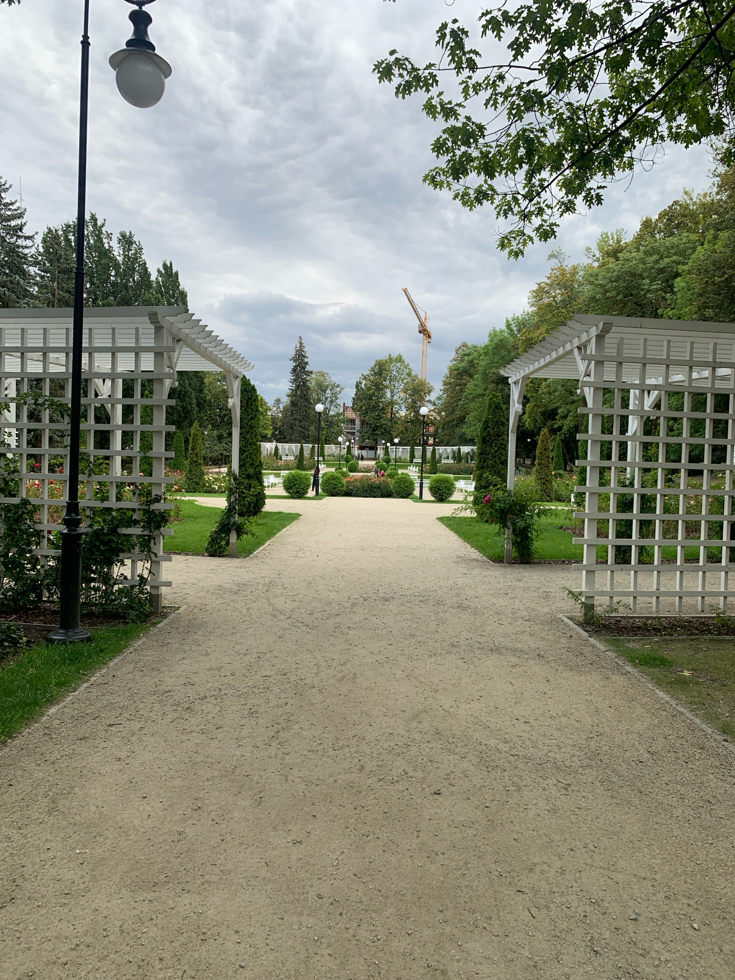 The image captures a view through white trellis archways in a park, likely Branicki Palace Garden in Białystok, Poland. The archways frame a gravel pathway leading into the park, with manicured lawns, hedges, and flowerbeds visible. A lone figure, possibly a woman in dark clothing, stands in the distance on the pathway. Beyond the park, a yellow construction crane is visible against a cloudy sky, suggesting ongoing development or renovation in the city. Trees, including conifers and deciduous varieties, surround the park area. A vintage-style lamppost stands to the left of the frame, near the foreground archway. The scene is characterized by orderly landscaping and hints of urban activity in the background.