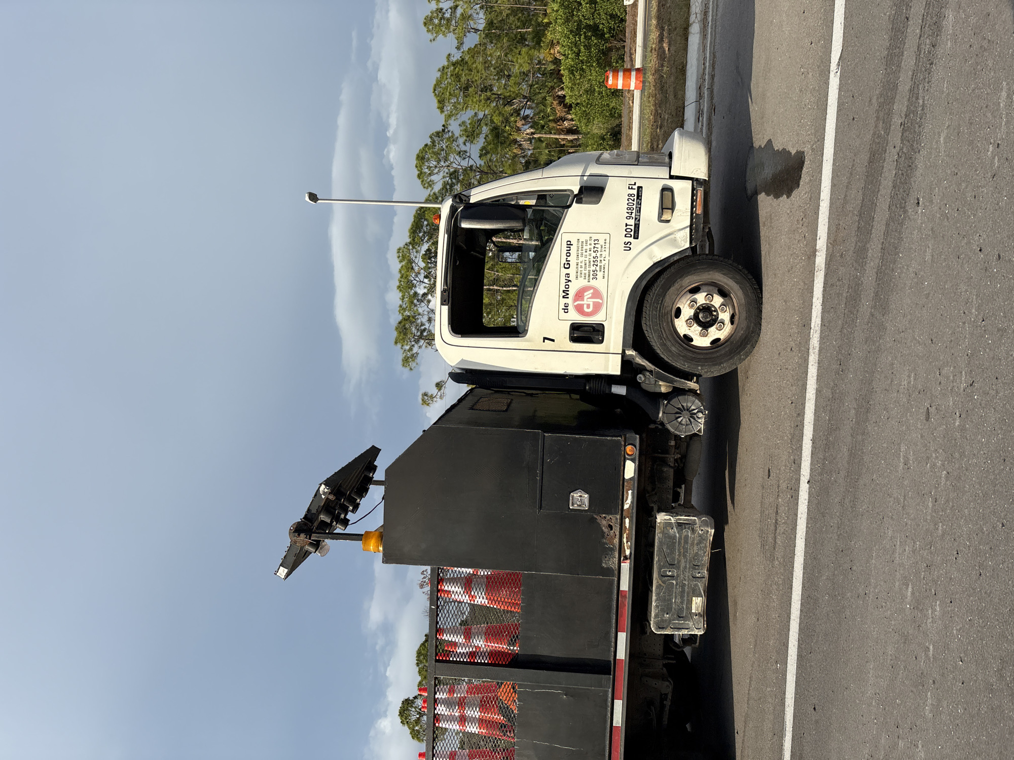 A white Isuzu truck with a black utility bed is parked on the right side of a two-lane road. The truck bed is loaded with orange and white traffic cones. A light is attached to the top of the truck bed. The truck has a "de Moya Group" logo on the passenger door. A US DOT number, 948028 FL, is visible on the truck's front fender. The background consists of green trees and a cloudy sky. An orange traffic barrel is visible on the side of the road. The scene is likely in Venice, Florida, United States.