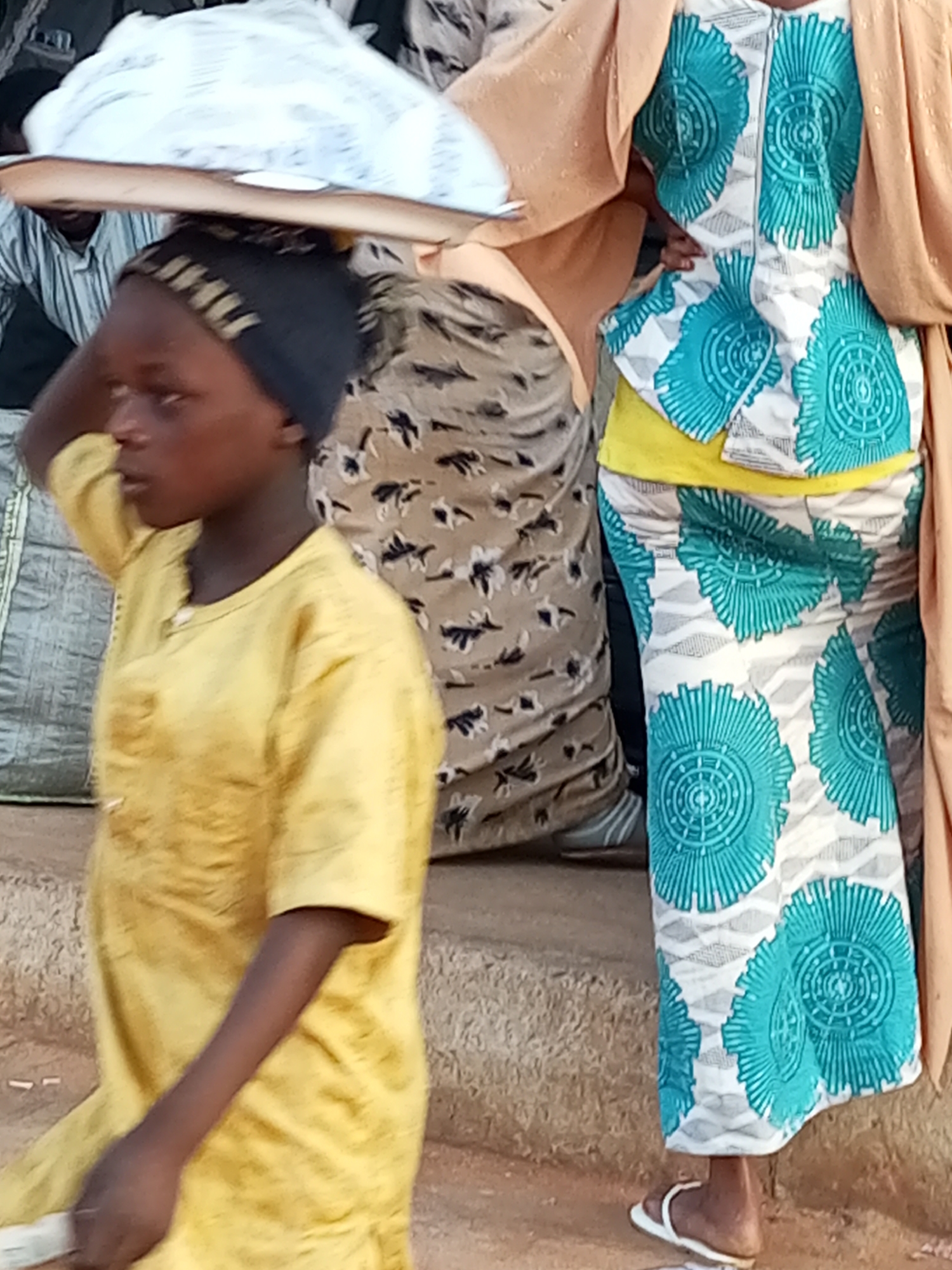 In Gombe, Nigeria, a young person is depicted in a medium shot from the side, carrying a large, flat object balanced on their head. They are wearing a yellow garment and a dark head covering. To their right, another person is visible, partially obscured, wearing a patterned outfit with teal and white circular designs, and a tan over-garment. This second individual is wearing white sandals. The background shows a textured, earthy surface, possibly a step or uneven ground, with some bags and other figures faintly visible. The overall scene suggests a market or public area.