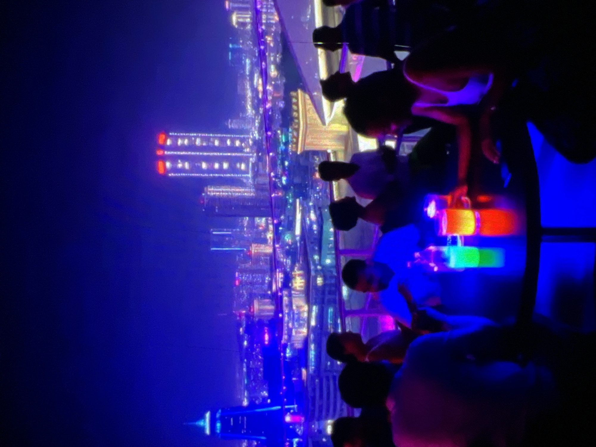 The image depicts a rooftop bar scene at night with a cityscape in the background, presumably in Lagos, Nigeria. Several individuals are present, some seated around a circular table and others standing, silhouetted against the vibrant city lights and the dark sky. The atmosphere is illuminated by neon and colored lights. On the table, there are two drinks, one appearing to be a brightly colored cocktail in a glass with a yellow and red glow, and another with a green glow in a bottle or glass. The city in the background features numerous tall buildings, some illuminated with lights, creating a dazzling urban panorama. The overall impression is one of a lively evening gathering at an upscale venue.