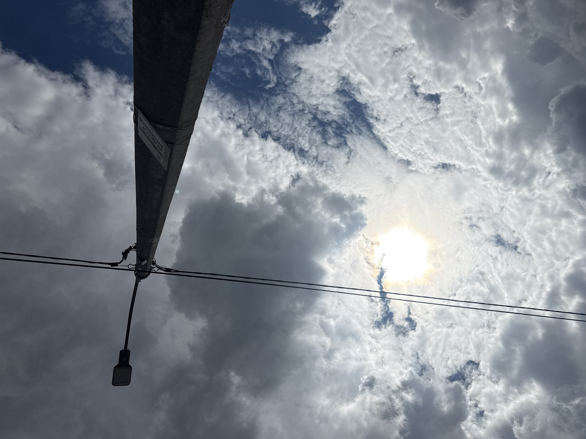 The image shows a view of the sky with clouds and a utility pole with a street light in Mytishchi, Russia. The sky is partially obscured by clouds, with the sun visible through a gap in the clouds. A gray concrete utility pole extends diagonally across the frame, with a street light fixture hanging from it. Two electrical wires run horizontally across the frame, intersecting the utility pole. A sign is affixed to the pole, though the text on it is not clearly legible. The overall scene presents an urban environment with a focus on infrastructure and weather conditions.
