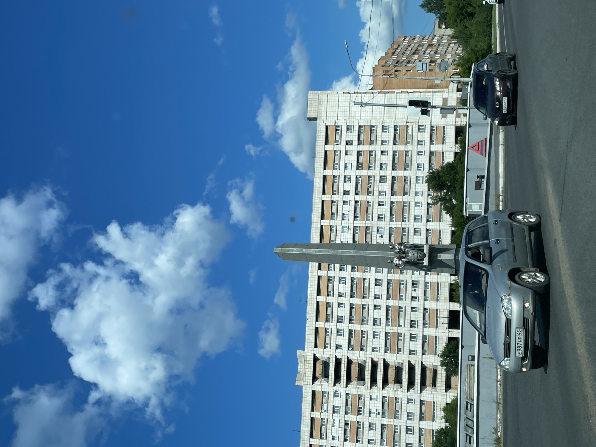 The image captures a bright, daytime scene on a street in Kirovo-Chepetsk, Russia. The sky is a vibrant blue, dotted with fluffy white clouds. In the background, a large, multi-story residential building with a light-colored facade and rows of windows dominates the skyline. To the right of this building, another, older brick building is visible. In the foreground, a wide road is present with several cars. A silver station wagon is prominently featured, its license plate reading "E 987 HP 43." To its right, a dark-colored sedan is also driving on the road. Between the buildings and the road, there appears to be a monument or obelisk with sculptures at its base. The overall environment suggests an urban setting on a clear day.