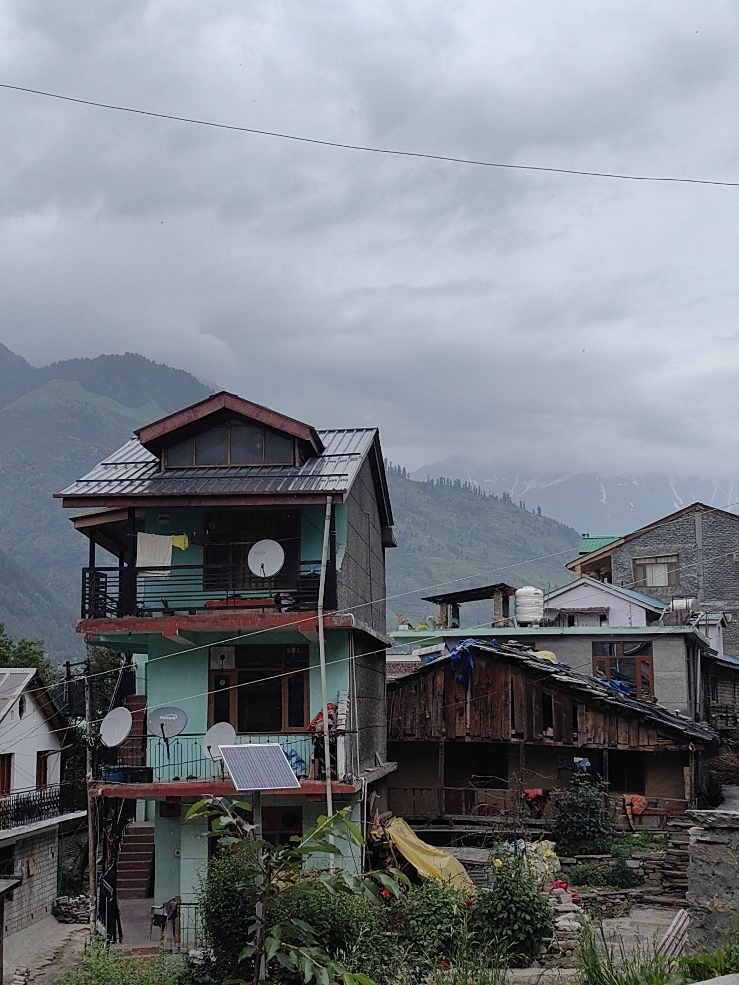 The image captures a view of buildings in Vashisht, India, against a backdrop of misty mountains under a cloudy sky. The foreground features a multi-story building painted turquoise and red, with balconies, satellite dishes, and a solar panel. Adjacent to it is a rustic wooden structure with a dilapidated roof and laundry hanging outside. Further buildings, some constructed of stone, are visible in the mid-ground and background. Lush green vegetation, including trees and bushes, is present throughout the scene. The overall atmosphere is overcast and damp, suggesting recent or ongoing precipitation.