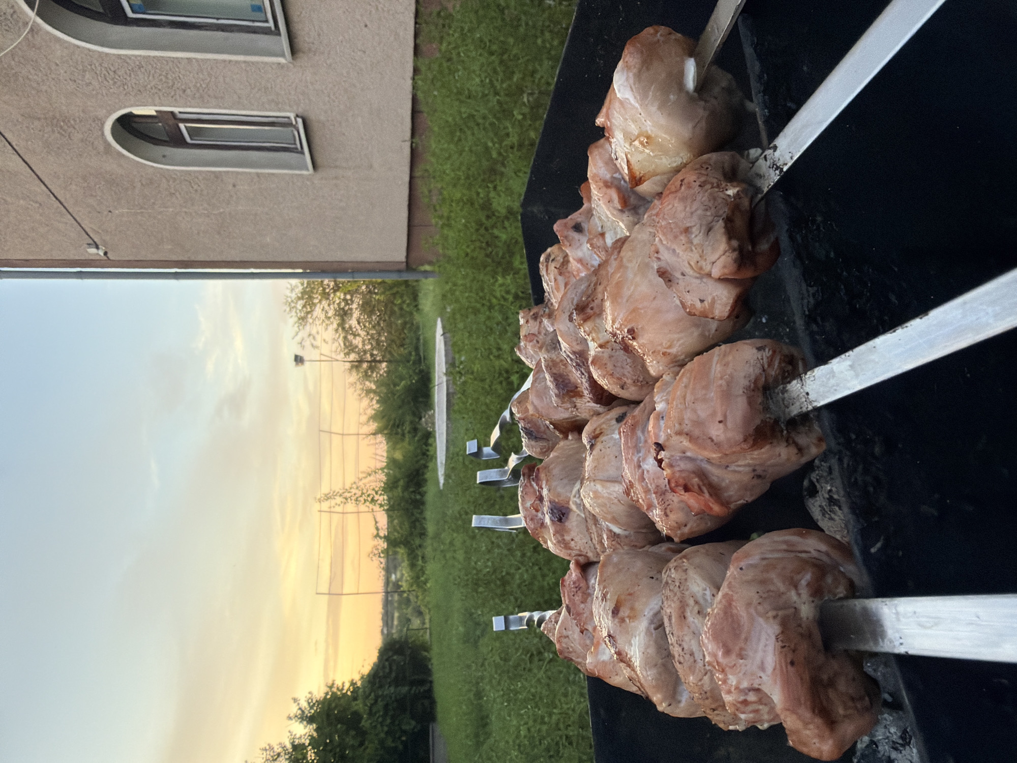 In Lozuvatka, Ukraine, a barbeque is in progress during sunset. Skewers of raw meat are arranged on a black grill, ready for cooking. The background features a grassy area, a building with arched windows, and a soft, orange-hued sky.
