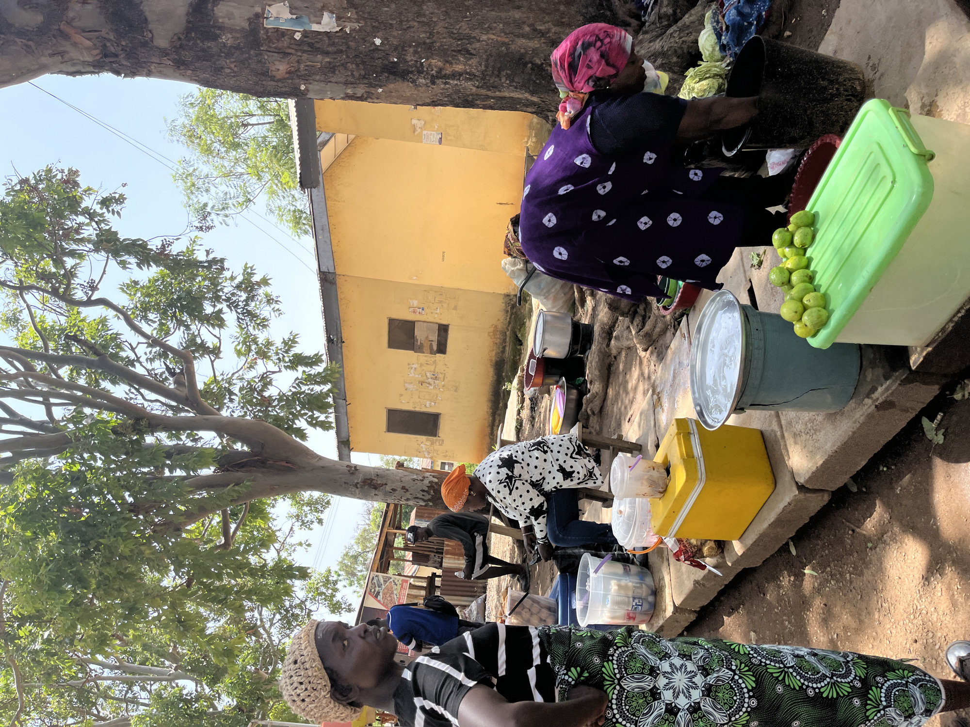 This is a daytime, eye-level, outdoor scene at a marketplace in Rafin Par, Nigeria. In the foreground, a woman wearing a patterned green and black skirt and a black and white striped top stands facing left, looking to her left. To her right, on a raised concrete ledge, are various containers including a yellow cooler box, several clear plastic buckets filled with bottles and other items, and a large turquoise bucket with a metal lid filled with water. In the middle ground, a woman wearing a white and black polka dot jacket and an orange headscarf is seated on a stool, looking down at her hands. Next to her are cooking pots and other market goods. Further back, a man sits on a stool. Behind the stalls, a yellow building with a corrugated metal roof is visible. A large tree trunk is on the right side of the frame. A second woman, wearing a purple cloth with white circular patterns and a pink headscarf, is bent over a black bucket on the right, appearing to be washing something. In front of her is a bright green cooler box with several green fruits on its lid. The ground is a dirt path, and overhead, branches and leaves from trees create dappled shade.