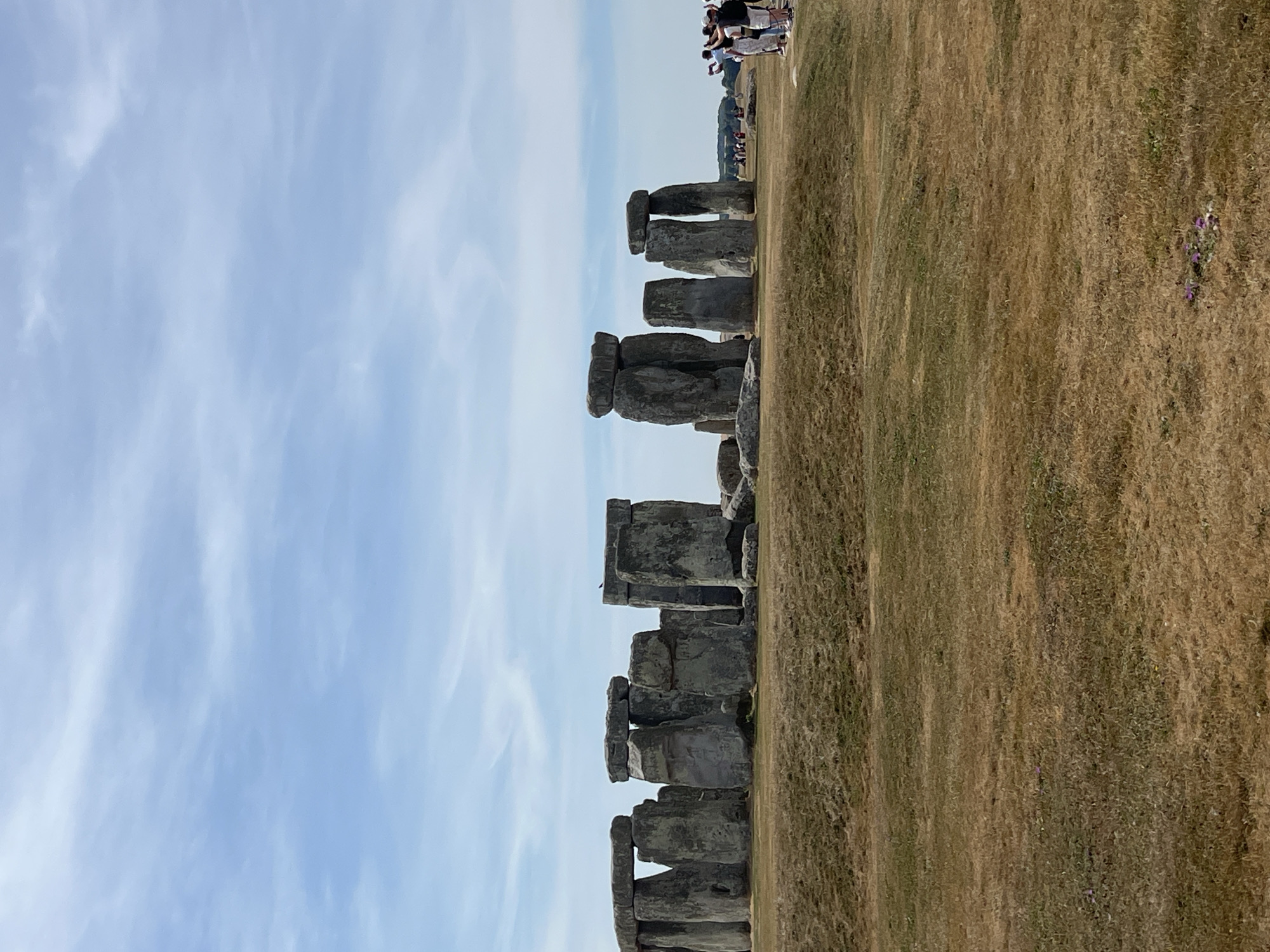 The image depicts Stonehenge, a prehistoric monument located near Amesbury, United Kingdom. The monument consists of a circle of standing stones, with some stones forming trilithons (two vertical stones supporting a horizontal stone). The stones are made of a greyish, rough-textured material. The surrounding landscape is dry, grassy terrain with some gentle undulations. In the background, a clear blue sky with scattered white clouds is visible. On the right side of the image, a group of people are standing on the grassy area, some of whom are holding up phones to take pictures. The lighting suggests it is daytime. The overall scene is natural and historical, with a focus on the ancient stone structure and its surroundings.