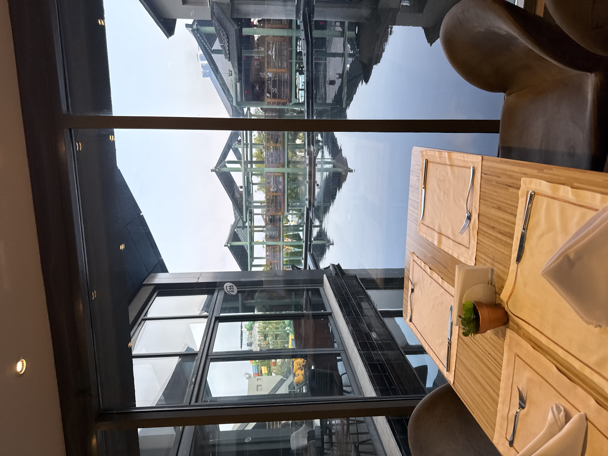 This image depicts a dining table set for two with placemats, napkins, forks, and knives, positioned in front of a large window overlooking a tranquil body of water. The water reflects the surrounding architecture, which features multi-level structures with open-air sections, wooden railings, and pitched roofs, reminiscent of a resort or a scenic dining establishment. Beyond the water, the sky is visible, appearing overcast or during twilight. The interior of the establishment shows glimpses of modern lighting and window frames. The scene appears to be within a hotel or a restaurant complex in the city of Aksu, Turkey, given the context. The atmosphere is peaceful and elegant, suggesting a fine dining experience.