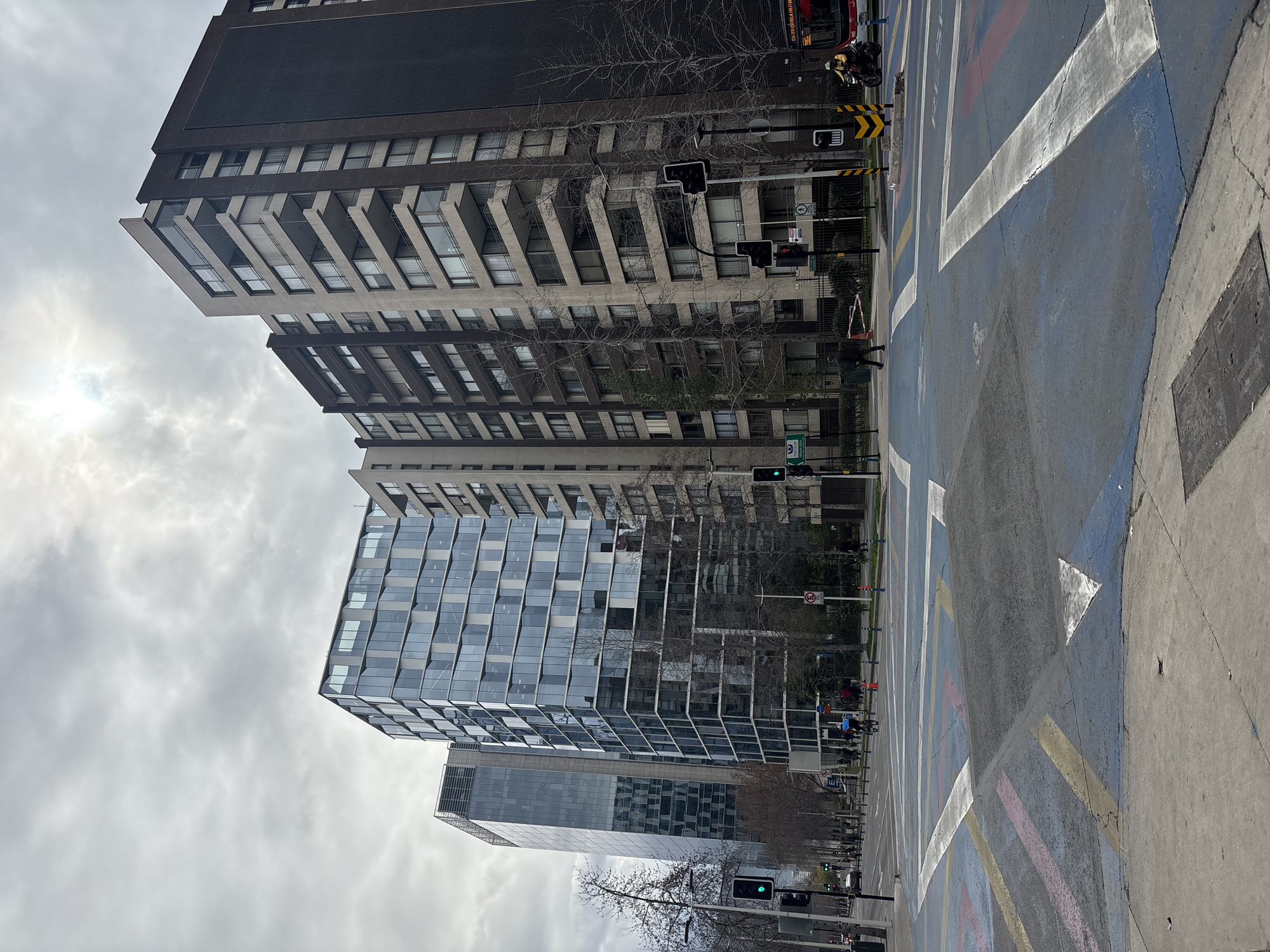 This is a street-level, medium-angle shot of a modern urban intersection in Providencia, Santiago, Chile. The scene is dominated by tall buildings, including a glass-fronted office building on the left and a darker, older apartment building on the right. The sky is overcast, with the sun visible through the clouds, creating a bright, diffused light.
In the foreground, the intersection features a complex network of painted lines and raised speed bumps, primarily in blue, white, and yellow, indicating pedestrian crossings and traffic lanes. Several traffic lights are visible, with the one on the far left showing a green light.
On the street, there are a few cars, a bus, and several pedestrians and cyclists. A person on a bicycle is cycling towards the right, and another pedestrian is walking across the street from left to right. A motorcycle with a rider wearing a yellow jacket is stopped at the intersection. Bare trees line the sidewalks, suggesting it might be winter or early spring. The overall impression is of a bustling but orderly urban environment.