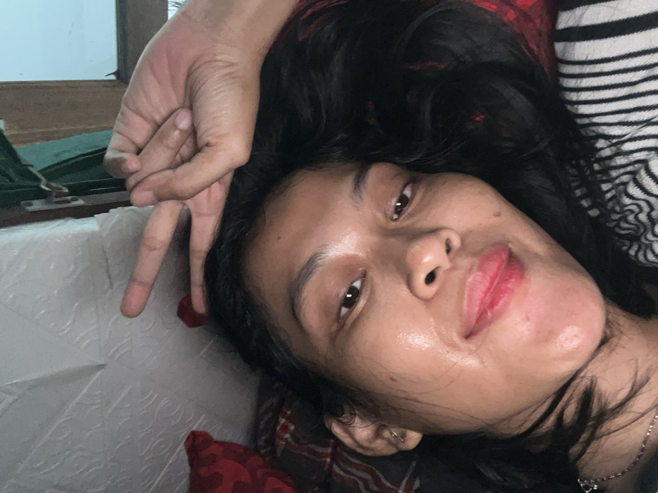 A woman lies on her side, facing the camera. Her black hair is tousled around her face. She has brown eyes and is wearing red lipstick. Her skin appears slightly shiny. A hand is positioned near her head, making a peace sign. She is lying on a bed or couch with a plaid blanket and a striped pillow. A window is visible in the upper left corner of the frame.
