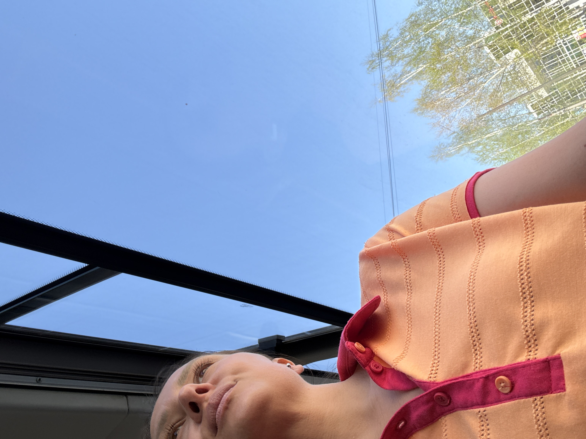 The image shows a woman lying down and looking up through a glass sunroof. Reflected in the glass are trees and buildings, which indicates an urban setting. The woman is wearing an orange and pink collared shirt. The glass sunroof is framed by black metal supports. Based on the location mentioned, the scene is likely in Chaikovsky, Russia.
