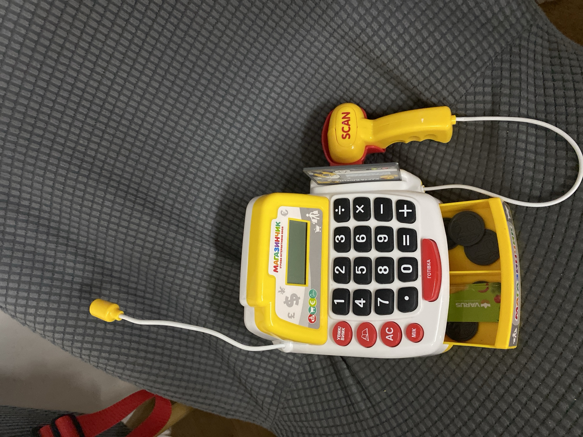 The image shows a toy cash register with a scanner accessory, placed on a grey, textured fabric surface. The cash register is predominantly white and yellow, with a yellow antenna extending from the top. It features a numerical keypad, various function buttons (including "AC", "УВМК/ВИМК", and "MIK"), and a small screen. A red drawer at the bottom contains play money and coins. The yellow scanner, connected to the cash register by a white cord, has the word "SCAN" printed on it. The background is out of focus, but appears to be a grey upholstered surface, possibly a chair or sofa. There are no people visible in the image. The items are arranged in a static, posed manner. The image was taken indoors.