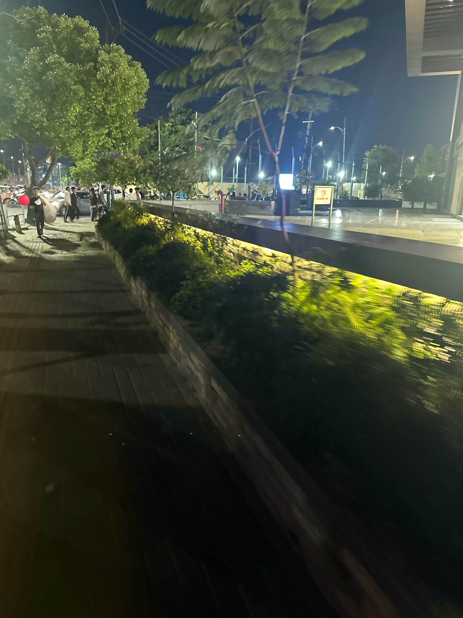 The image depicts a nighttime scene along a sidewalk in Kathmandu Metropolitan City, Nepal. The sidewalk is paved with rectangular stones and is illuminated by artificial lights, casting shadows. To the right of the sidewalk, a raised planter box filled with dense green foliage is visible, also lit from above. Beyond the planter box, a modern building facade with large glass windows can be seen. Further into the background, streetlights illuminate a paved area that appears to be a walkway or plaza. Several trees are present, with lush green leaves. A group of people, appearing to be young adults, are walking and standing on the sidewalk and in the background. Some are carrying bags, and one individual is walking alone on the sidewalk in the foreground. Cars are parked or moving on a street to the left of the sidewalk. The overall atmosphere is one of an urban environment at night, with activity and illumination.