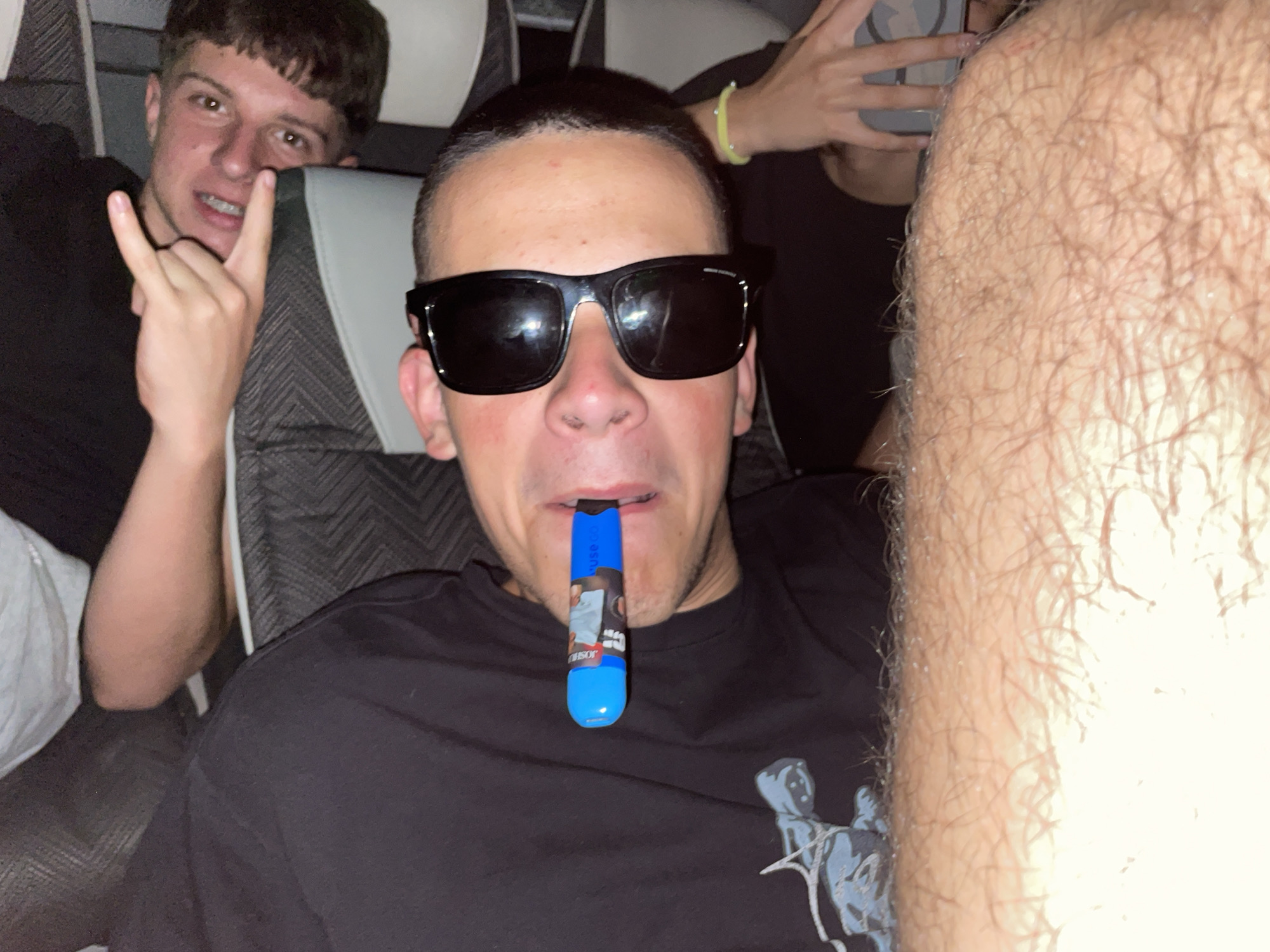 The image shows three people seated in a vehicle. One person is visible in the background, holding up a "rock on" hand gesture. The second person, in the foreground, is wearing sunglasses and has a blue vaping device in their mouth. The third person has an arm in the foreground, covered in dense hair. The location is unknown.