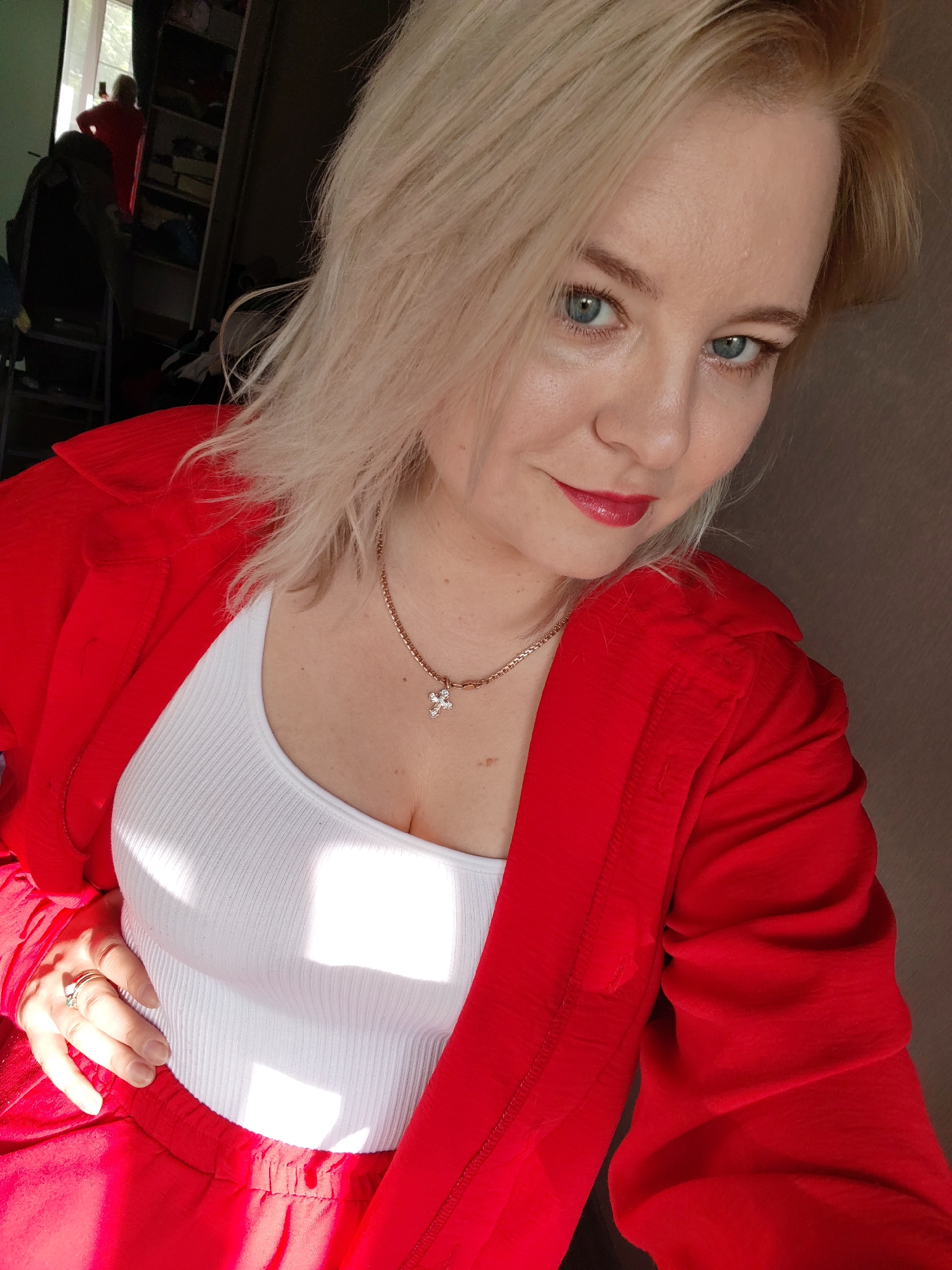 A fair-skinned woman with blonde hair and blue eyes is posing for a selfie indoors. She's wearing a white ribbed tank top and a bright red button-up shirt, along with a gold chain necklace with a cross pendant. Her lips are red, and she's wearing a ring on her finger.
In the blurry background, what appears to be a closet or shelving unit is visible. A person in a red garment is near a window. The location is unknown, and there is no clear indication that this is in Izhevsk, Russia.