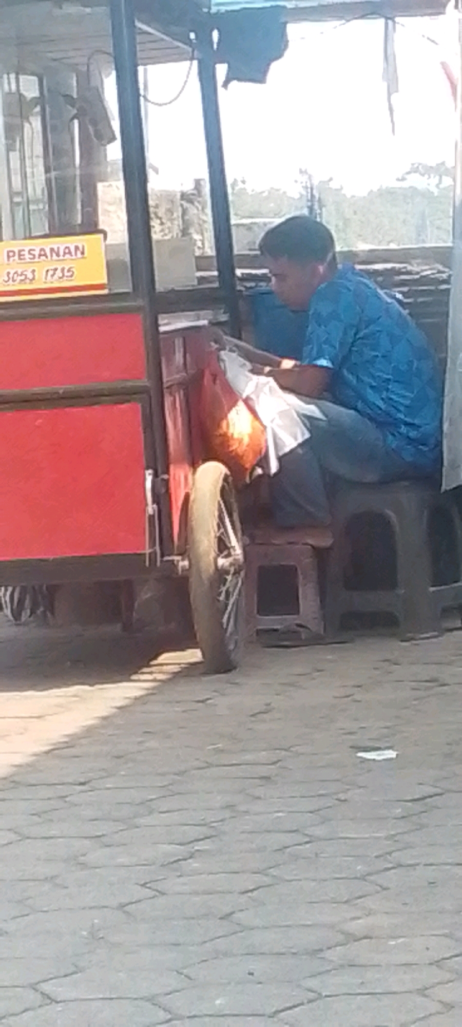 A man in a blue patterned shirt and dark pants is seated on a black stool next to a red cart. The cart has a yellow sign that reads "PESANAN" with "3053 1735" below it. The man is holding an orange bag and appears to be looking down at it. The cart has a visible wheel on the left side. The ground is paved with hexagonal stones, and there are shadows cast on the ground. The background shows a bright sky and some foliage. The location is identified as Sirnagalih, Indonesia.