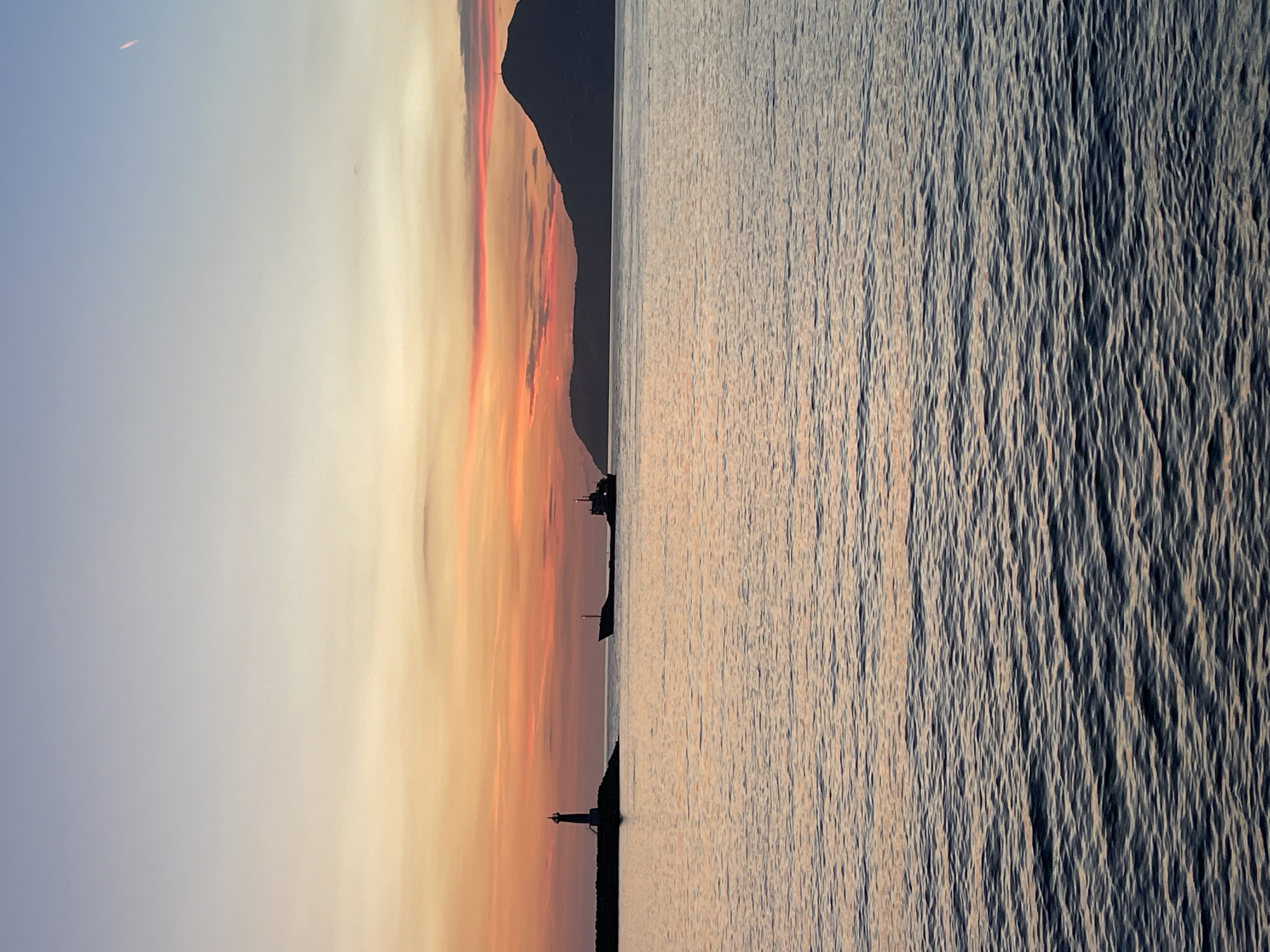 This image depicts a scenic view of the sea and sky at sunset, with a dark, silhouetted mountain range in the background. The sky is a gradient of soft blues and vibrant oranges, indicative of a sunset or sunrise. There are several lighthouses visible along the coastline, standing as solitary structures against the dramatic sky and sea. The sea itself is calm, with gentle ripples reflecting the colors of the sky, creating a peaceful and serene atmosphere. The image captures the beauty of nature and the tranquility of the coast. There are no visible people or text in the image. The location is Bar, Montenegro.