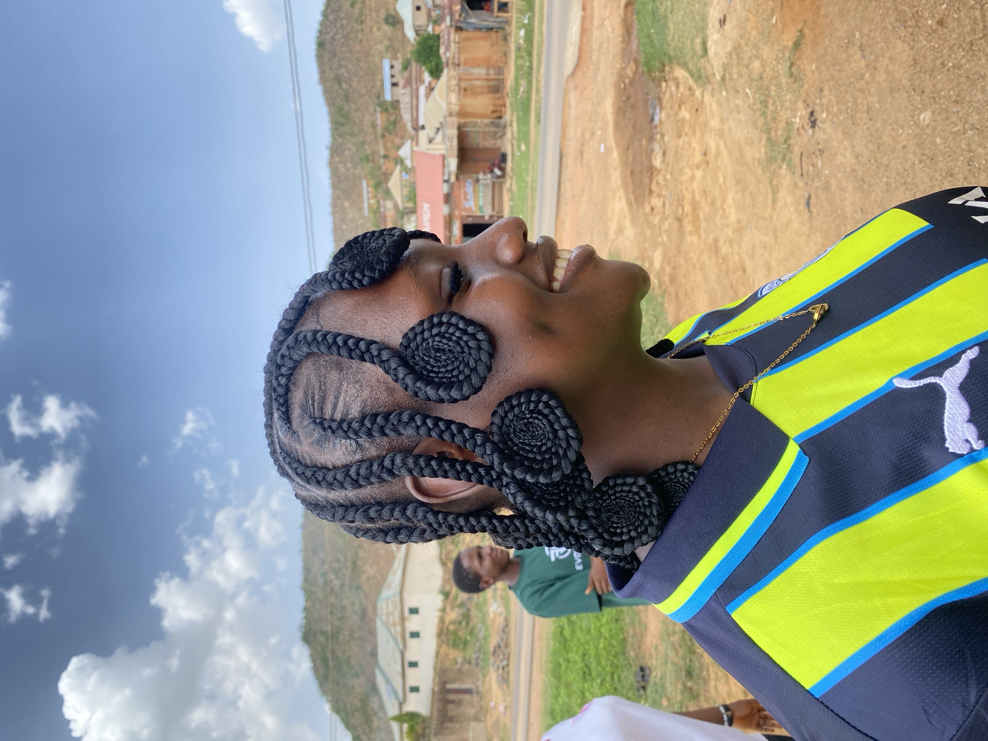 A woman is the primary subject, seen in a profile shot with her face tilted upward, smiling and eyes closed, in an outdoor setting. She has dark skin and her hair is styled in intricate braids, some coiled into spiral patterns. She wears a black and neon yellow Puma sports shirt with blue stripes.
The background reveals a landscape with buildings and hills. The location is in Gwong, Nigeria, with a dirt road and visible buildings in the distance. The sky is partly cloudy. In the mid-ground, there is a man in a green shirt and part of another person's white shirt visible in the foreground.