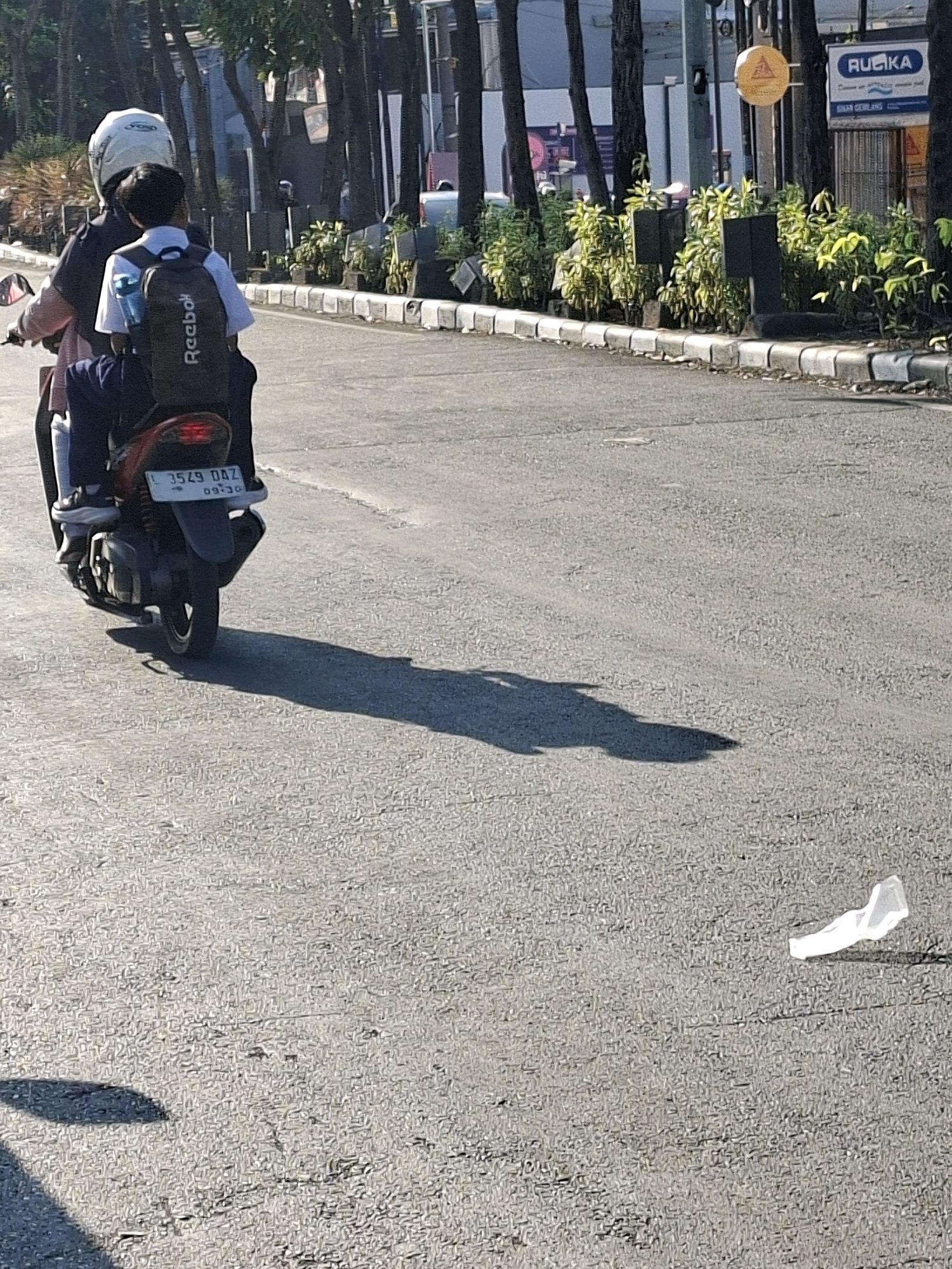 The image depicts two individuals riding a motorcycle on a sunlit asphalt road in Surabaya, Indonesia. The person in front wears a white helmet and a pinkish jacket, while the person behind, likely a child, wears a white shirt and a backpack with "Reebok" written on it. The motorcycle has a license plate visible, reading "L 3549 DAZ". The background shows a row of trees lining the street, with buildings and signs visible further in the distance, including one with the text "RUIKKA". The road surface is textured and shows tire marks, with a noticeable shadow cast by the motorcycle and its riders. A crumpled piece of white paper is visible on the road to the right of the frame. The lighting suggests it is daytime, with strong sunlight creating distinct shadows.