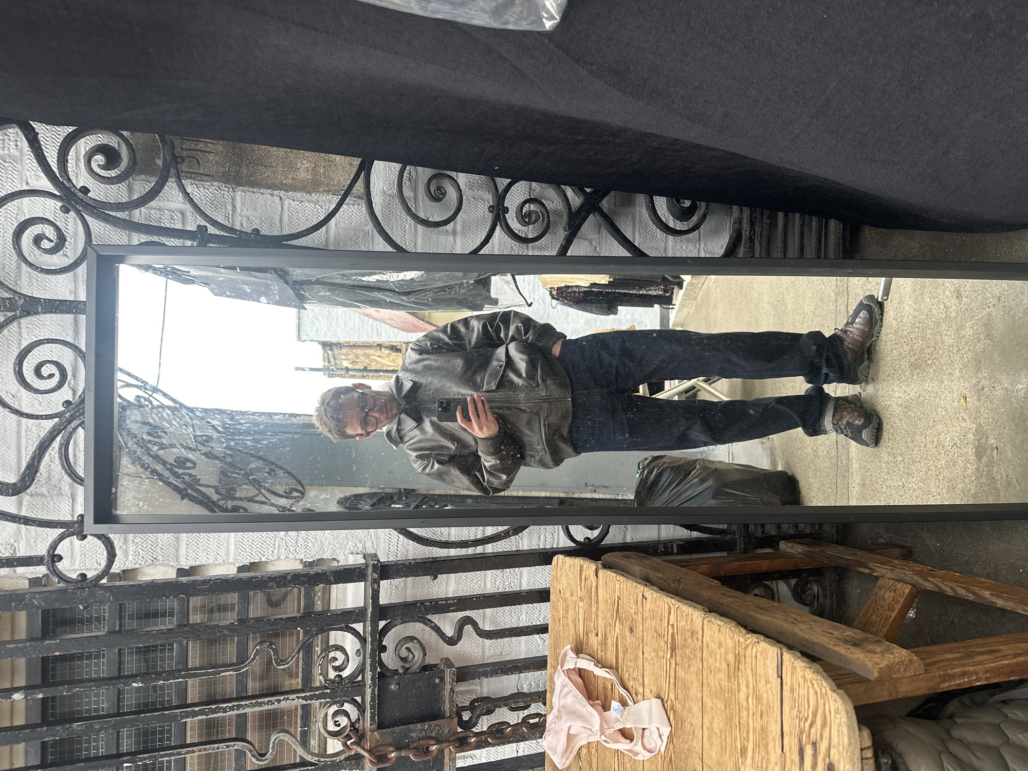 The image shows a person in a mirror standing on the street in London, UK. The person is wearing a leather jacket, jeans, and hiking boots, and is holding a mobile phone. The mirror reflects the surroundings, including buildings, wrought iron fences, a garbage bag, and a wooden table with a light pink bra on it. The sky is overcast.