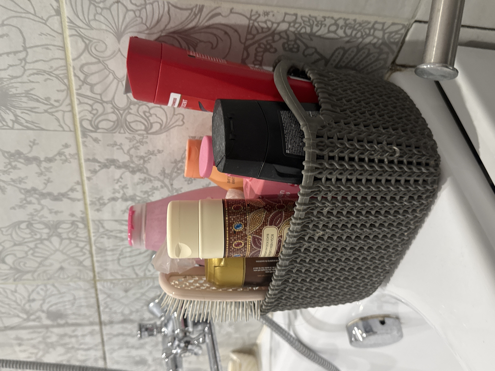 The image shows a gray woven plastic basket filled with various toiletries on the edge of a white bathtub. The basket contains a pink bottle, a small yellow bottle, a dark bottle with a patterned label, a pink bottle, an orange bottle, a black bottle with a gray lid, and a red bottle. A light-colored hairbrush is also partially visible in the basket. The background consists of white tiles with a gray floral pattern. A chrome faucet and showerhead are visible to the left. The scene appears to be a bathroom. The provided metadata indicates the image was taken in Svitlovodsk, Ukraine.