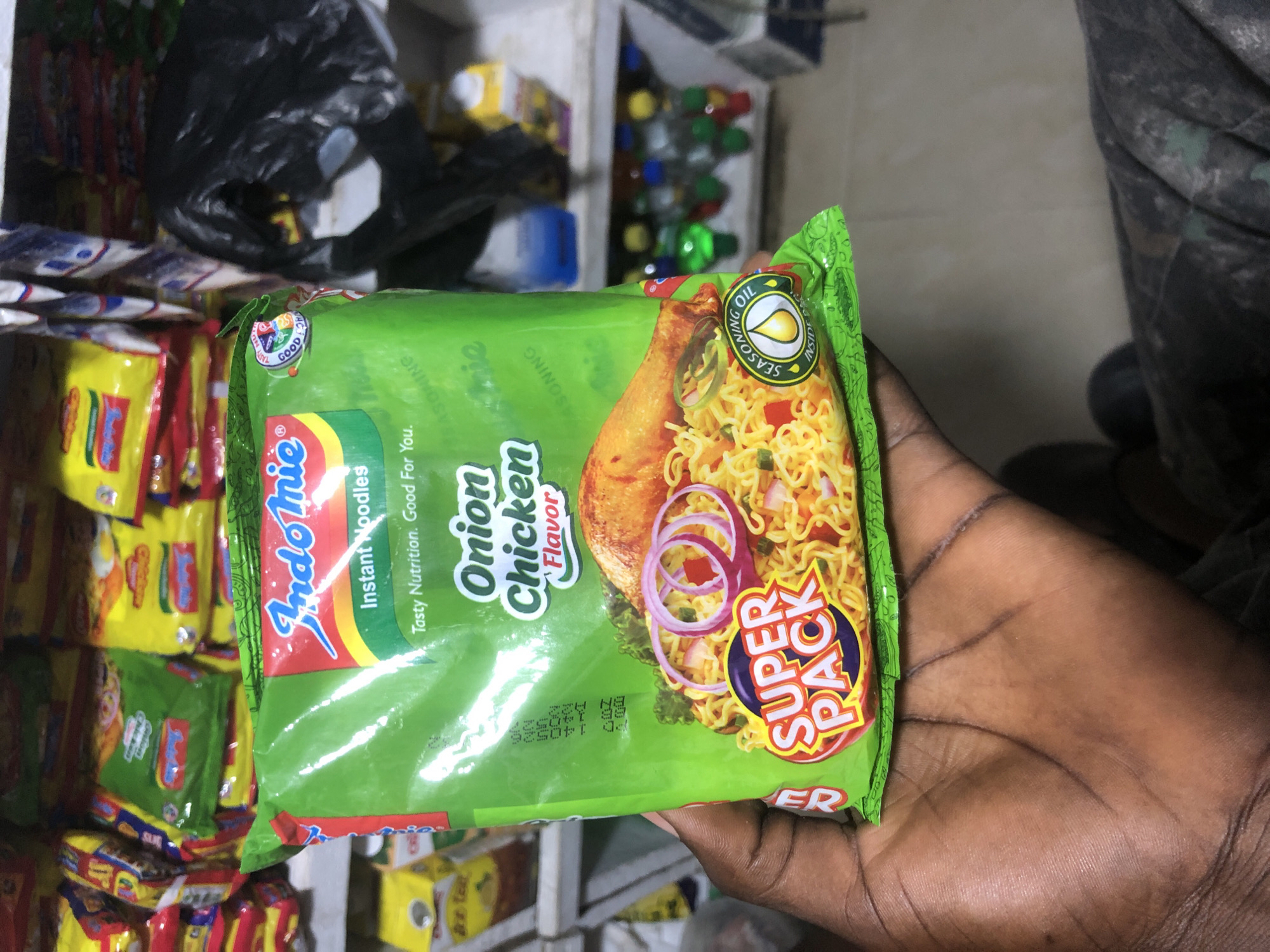 In Ado, Nigeria, a person's hand holds a green packet of Indomie Onion Chicken flavor instant noodles. The packet is a "Super Pack" and features an image of cooked noodles with chicken, onions, and seasoning. The background shows shelves stocked with more noodle packets, plastic bags, and bottles, suggesting the setting is a shop or market stall. The lighting is moderate, indicating an indoor environment.