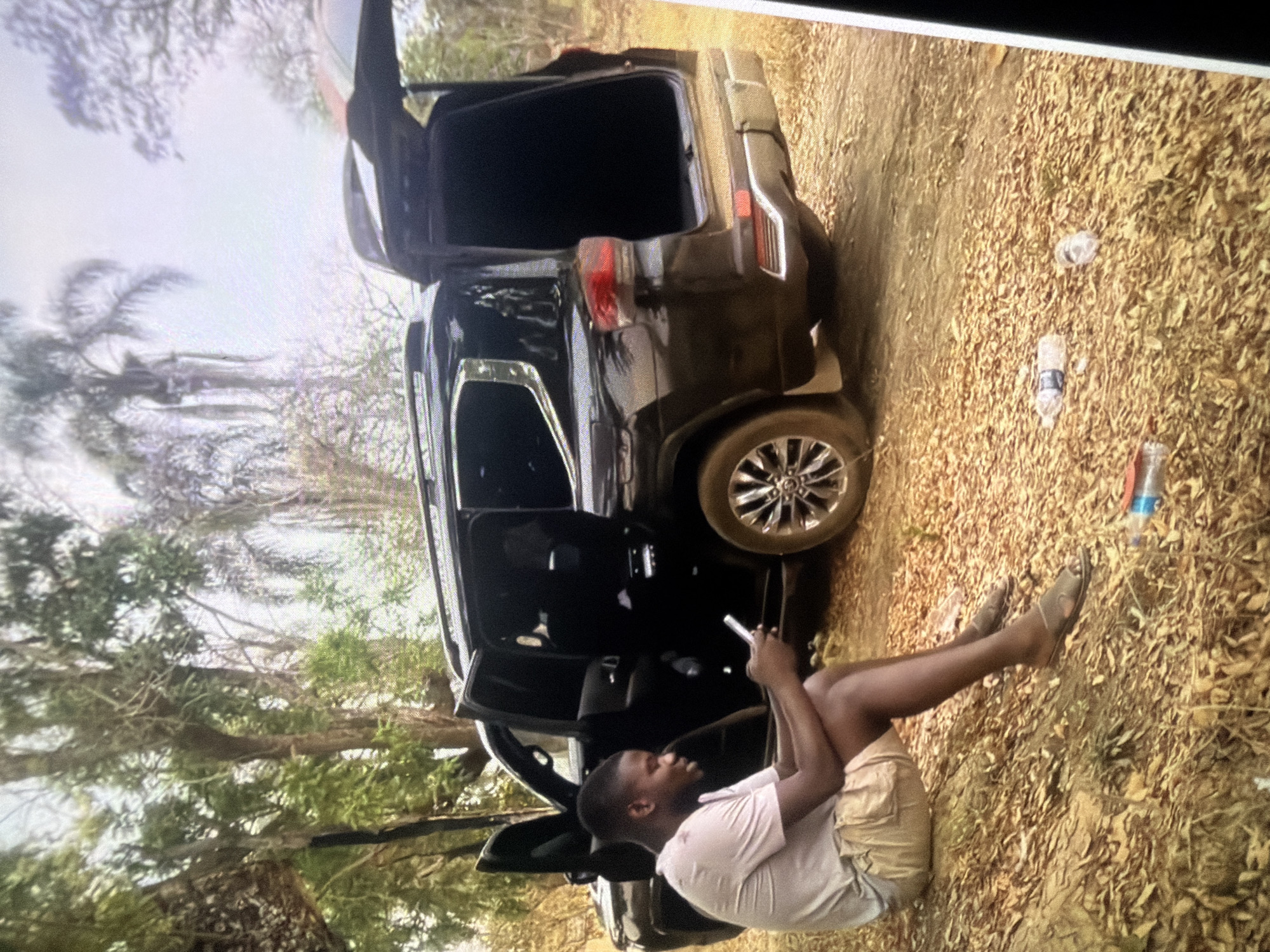 A young man sits on the ground next to a black SUV with its doors and trunk open. The man is wearing a light-colored t-shirt and khaki shorts, and he is looking at his phone. The SUV is parked on a dirt road in a wooded area. The ground is covered in dry leaves and branches. Two empty plastic water bottles are visible on the ground near the SUV. The location is identified as the city of Bukuru, Nigeria.
