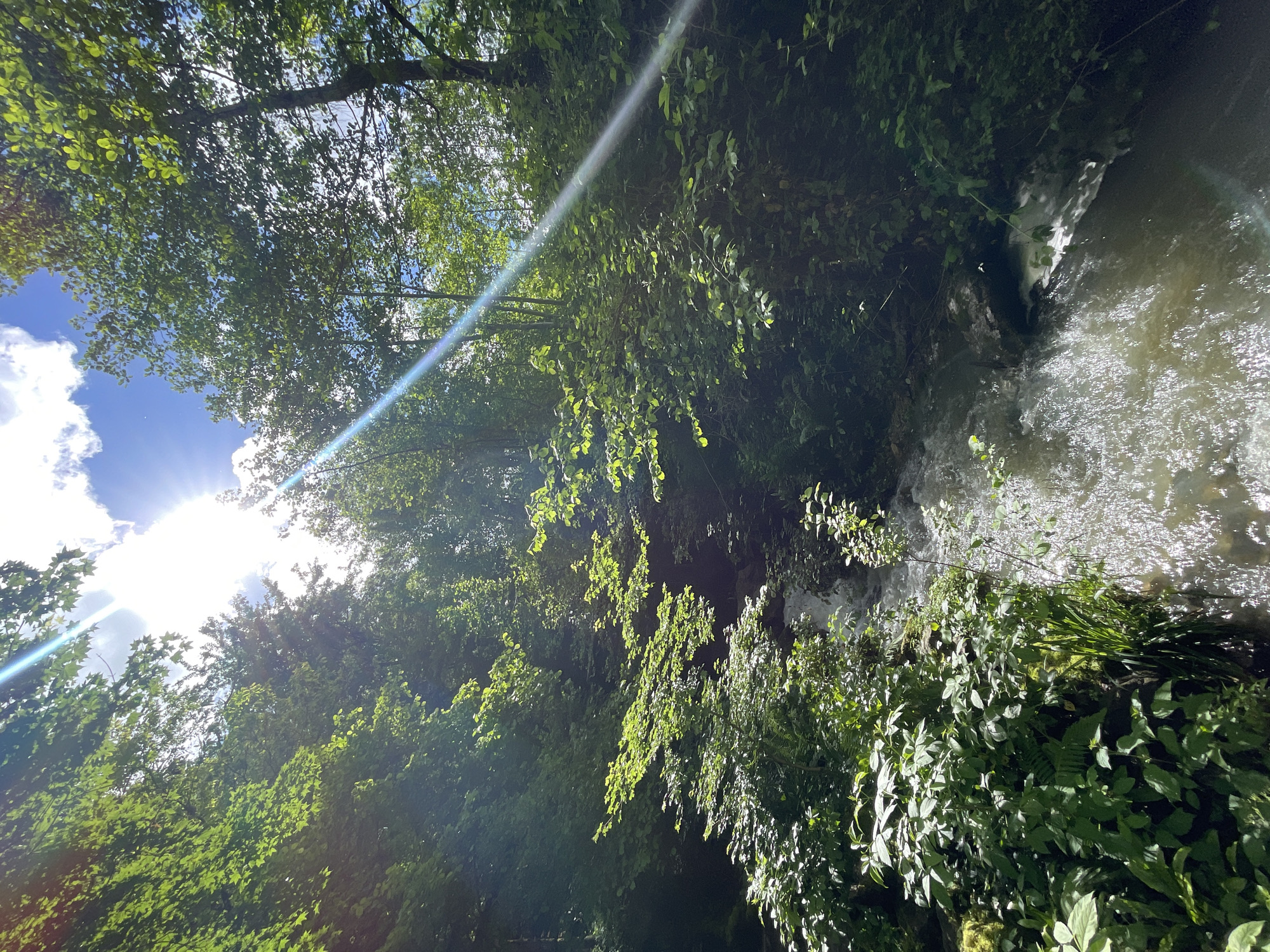 The image depicts a natural scene in Solokh-Aul, Russia. It features a waterfall cascading down a rocky cliff face covered in lush green vegetation. The trees above filter sunlight, creating dappled light patterns on the foliage. The water is turbulent and foamy as it flows down the waterfall. The sky is visible through the canopy of trees, showing blue sky and white clouds. A lens flare from the sun is visible in the upper part of the image.