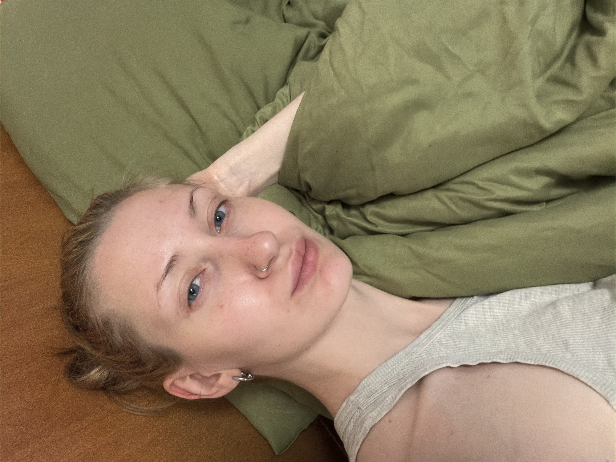 A young woman with blonde hair pulled back into a bun is lying in bed, looking at the camera. She is wearing a gray tank top and has silver hoop earrings and a nose ring. A green comforter is pulled up around her. The background consists of a wooden headboard and a green pillow. There is no indication of the location being Saint Petersburg, Russia.