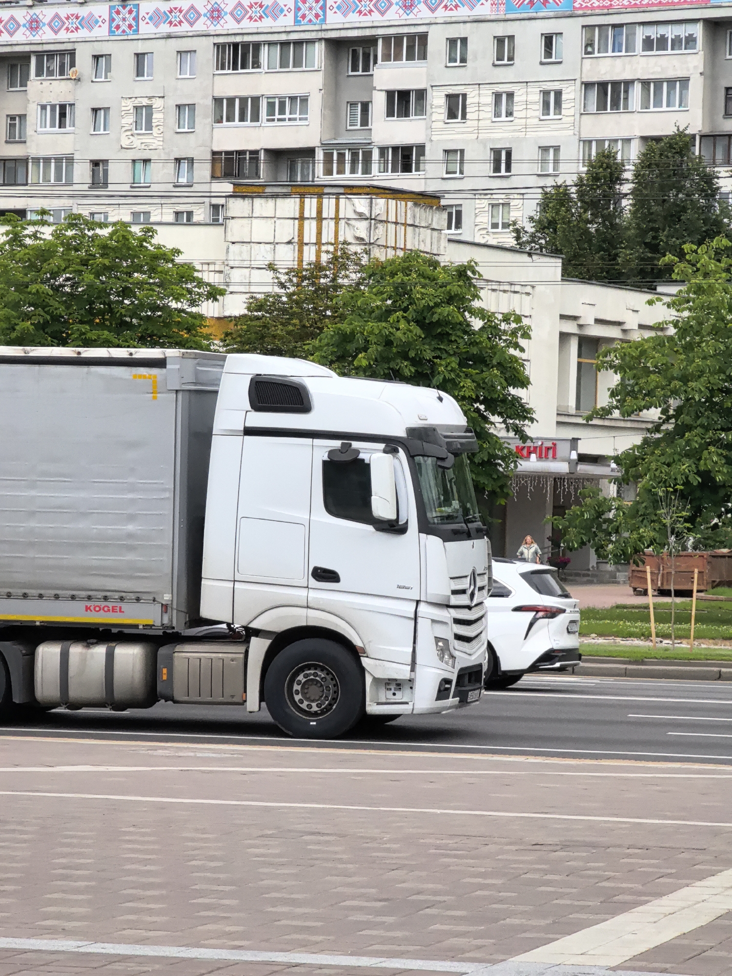 An exterior, medium shot captures a white truck with a silver trailer driving on a street in an urban area. The truck is a Mercedes-Benz Actros. The trailer is branded with the word "KOGEL." In the background, a white building with several windows and a decorative pattern along the top is visible. Trees are scattered throughout the scene. A white car is parked behind the truck on the right side of the frame. A woman is standing near the entrance of the building. The street is paved with asphalt and marked with white lines. The foreground consists of a sidewalk made of interlocking bricks.