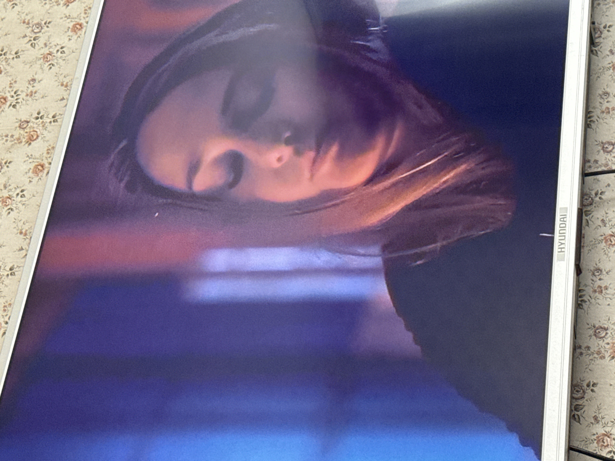 A close-up of a Hyundai television screen displaying a woman's face. The background is a blurred mix of red, purple, and blue hues. The television is on a surface with a floral pattern. The location is Mytishchi, Russia.