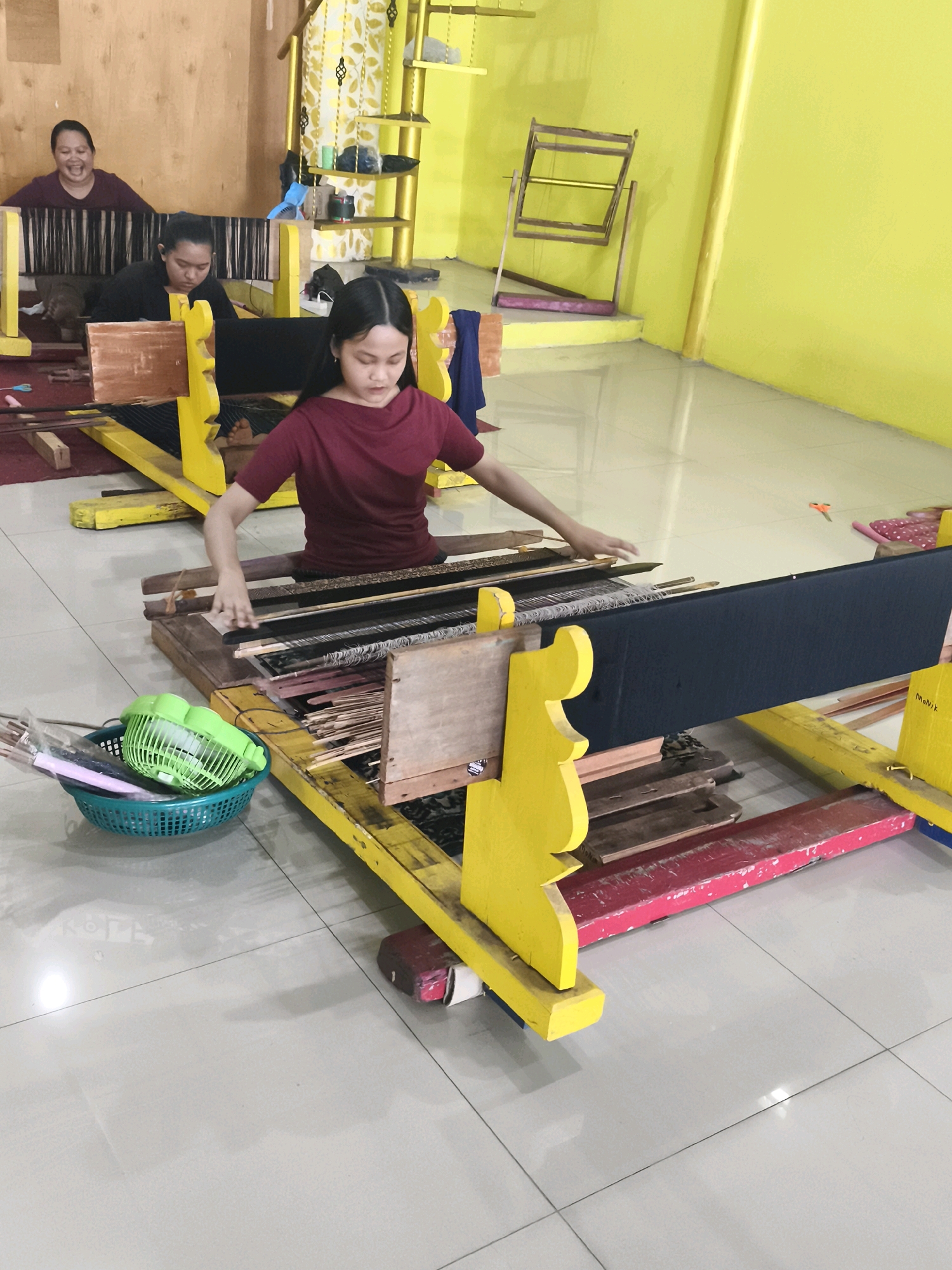 The image shows a group of Indonesian women working on a weaving project, likely in Palembang, Indonesia, given the distinct yellow and red looms and the presence of traditional Indonesian crafts. In the foreground, a young woman in a maroon shirt is actively engaged in weaving, her hands skillfully manipulating threads on a large loom. Behind her, another woman in a black shirt works on a similar loom, and further in the background, a third woman in a maroon shirt is visible, also participating in the activity. The environment appears to be a workshop or studio, with the characteristic bright yellow walls of Palembang's artisanal spaces and wooden accents. The looms themselves are brightly colored, a common feature in local craft workshops. The floor is tiled, reflecting the bright lighting. A green basket filled with weaving tools sits near the foreground loom, suggesting a busy and productive workspace. The overall impression is one of traditional craftsmanship and community collaboration.