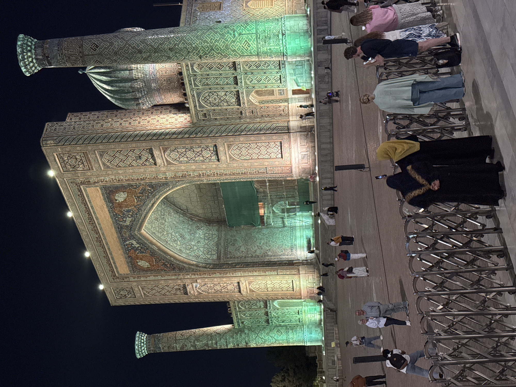 The image shows a nighttime view of the Registan Square in Samarkand City, Uzbekistan. The focus is on the illuminated facade of a large building, featuring intricate tilework and architectural details. Two tall, ornate pillars flank the building. A crowd of tourists stand near a metal fence and some are taking photos.